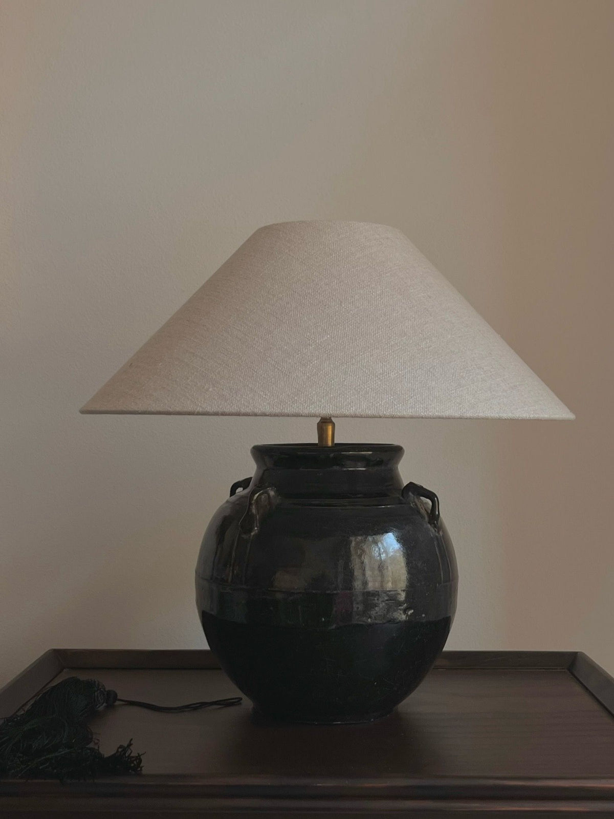 The Médecine Antique Glazed Lamp with Ears, featuring a wide cream shade and glossy dark ceramic base with handle-like protrusions, rests on a dark wooden surface against a plain light-colored wall.