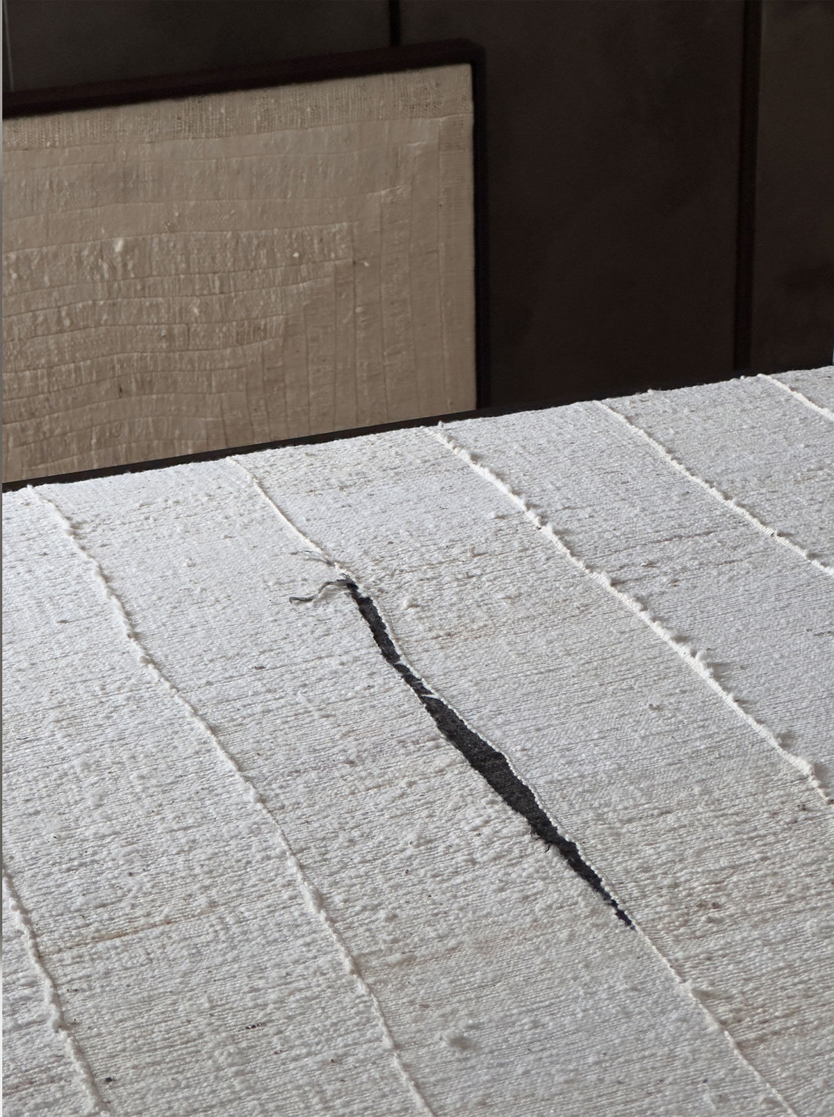 A detailed view of the "Mast, 2024" artwork by Bec Kirby reveals a visible crack in its textured, white, ridged surface that resembles plaster or stucco. In the background, a blurred rectangular object subtly contrasts with a framed segment of solid oak.