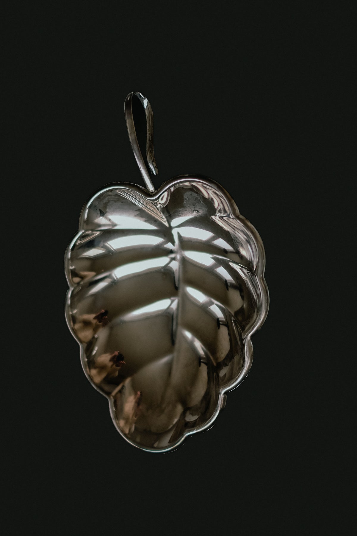 Set against a dark background, the "Serving Dish Leaf" by septembre studios is a reflective, metallic object with a small top loop. Its smooth, shiny surface and detailed vein patterns exude vintage charm and timeless elegance.