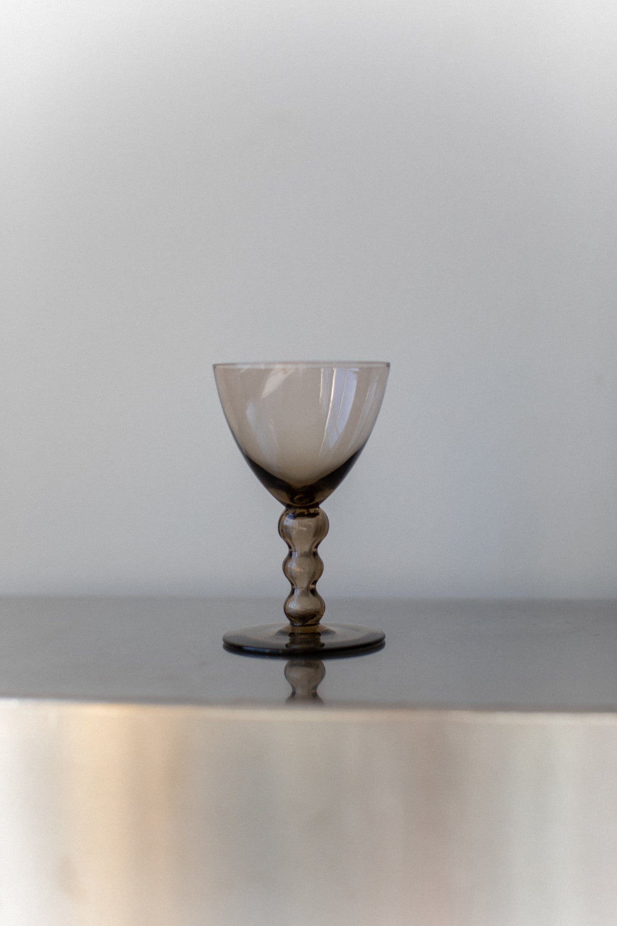 An Art Deco Glass from Out For Lunch features smoky gray glass resting on a reflective surface, enhancing the drinking experience. Its sculpted, curvy stem and wide bowl exude sophistication against a soft, neutral background.