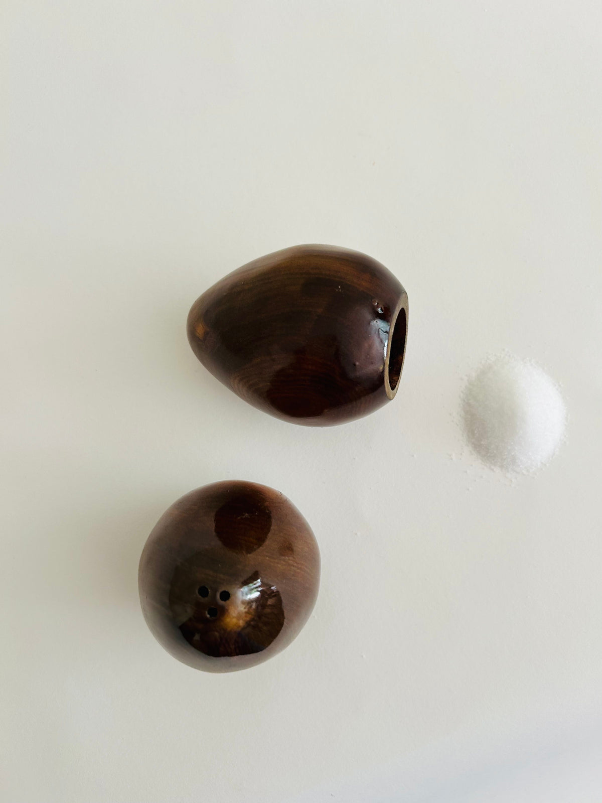 The Vintage Danish Wooden Salt & Pepper Set by Flock Studio features two glossy shakers in the spirit of mid-century modern design, elegantly displayed on a white surface. One shaker lies on its side next to a small pile of salt, while the other stands upright with multiple holes visible. This set is a perfect addition to any vintage tableware collection.