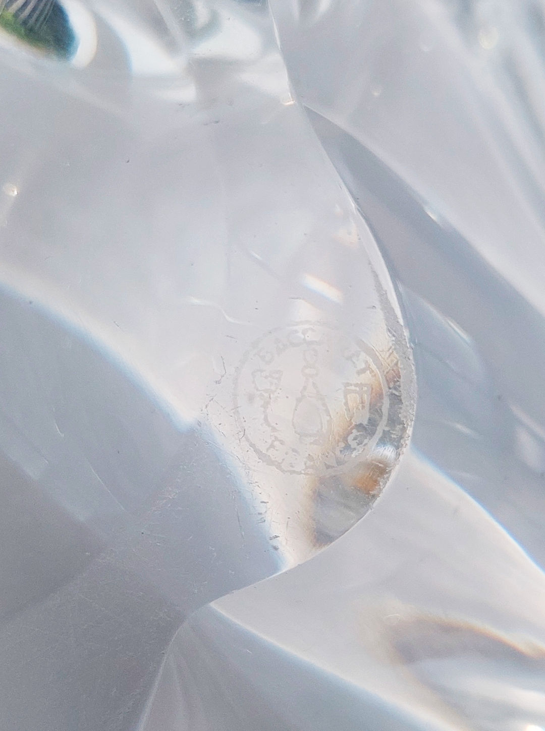 A detailed surface showcases an embossed circular logo with artistic flair, akin to the Les Objoies Crystal Baccarat Cup. Encircling small text surrounds a central design, its details subtly harmonizing with the crystal-like elegance of the texture.