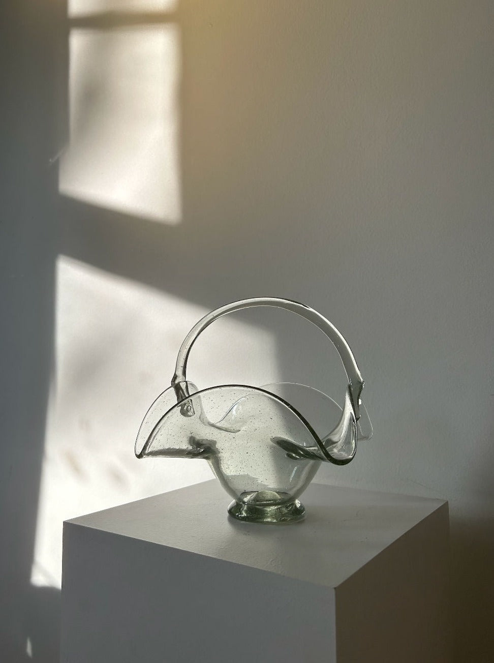 A clear glass, abstract-shaped sculpture, reminiscent of a 70's Vintage Glass Basket by Les Objets Amsterdam, sits on a white pedestal against a plain wall. Sunlight filters through a window, casting soft shadows and highlighting the delicate curves and thick quality glass of the piece.