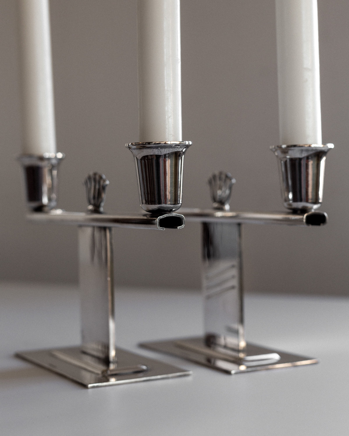 A close-up of a pair of Spigel Silver-Plated Art Deco Candle Holders, each featuring three elegant tall white candles. The sleek and minimalist design highlights Scandinavian craftsmanship with its reflective surface. They are arranged on a light-toned surface with a neutral backdrop.