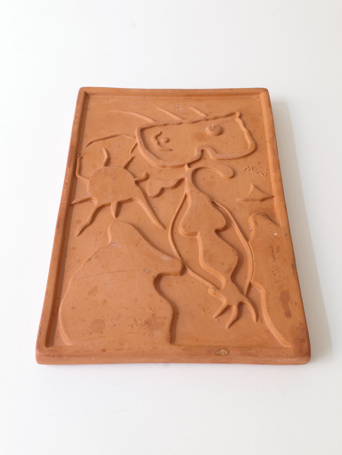 The Terracotta Relief Plaque Signed Joan Miró (1938) by Collection Apart features an abstract figure with elongated limbs and a bulbous head against a sunlit backdrop, set on an earth-toned rectangular slab, capturing the whimsical style of the Catalan surrealist artist.