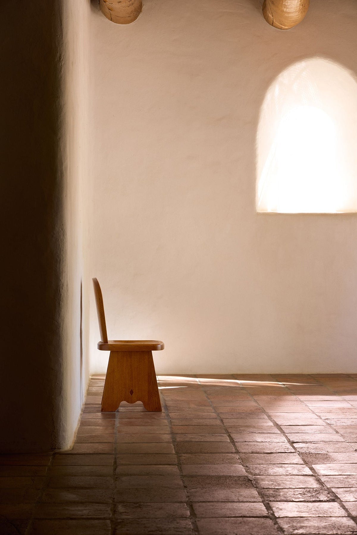 A Rambling Chair in Natural French Oak Wood by Lemon, reminiscent of a milk stool in its utilitarian functionality, is placed against a white, textured wall in a minimalist room. Sunlight filters through an arched window, casting a soft glow on the brick-tiled floor, creating a serene and peaceful ambiance.