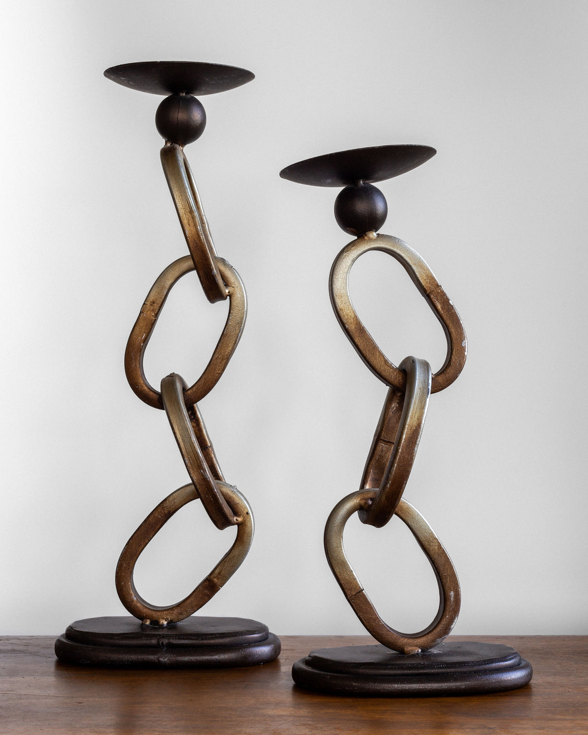 The "Postmodern Sculpture Duo" by Spigel features two metallic candle holders with a chain link design, standing on a wooden surface against a plain background. The chains are twisted and boast an antique brass finish, supporting round candle cups at the top. With varying heights, these holders add visual interest to any industrial decor setting.
