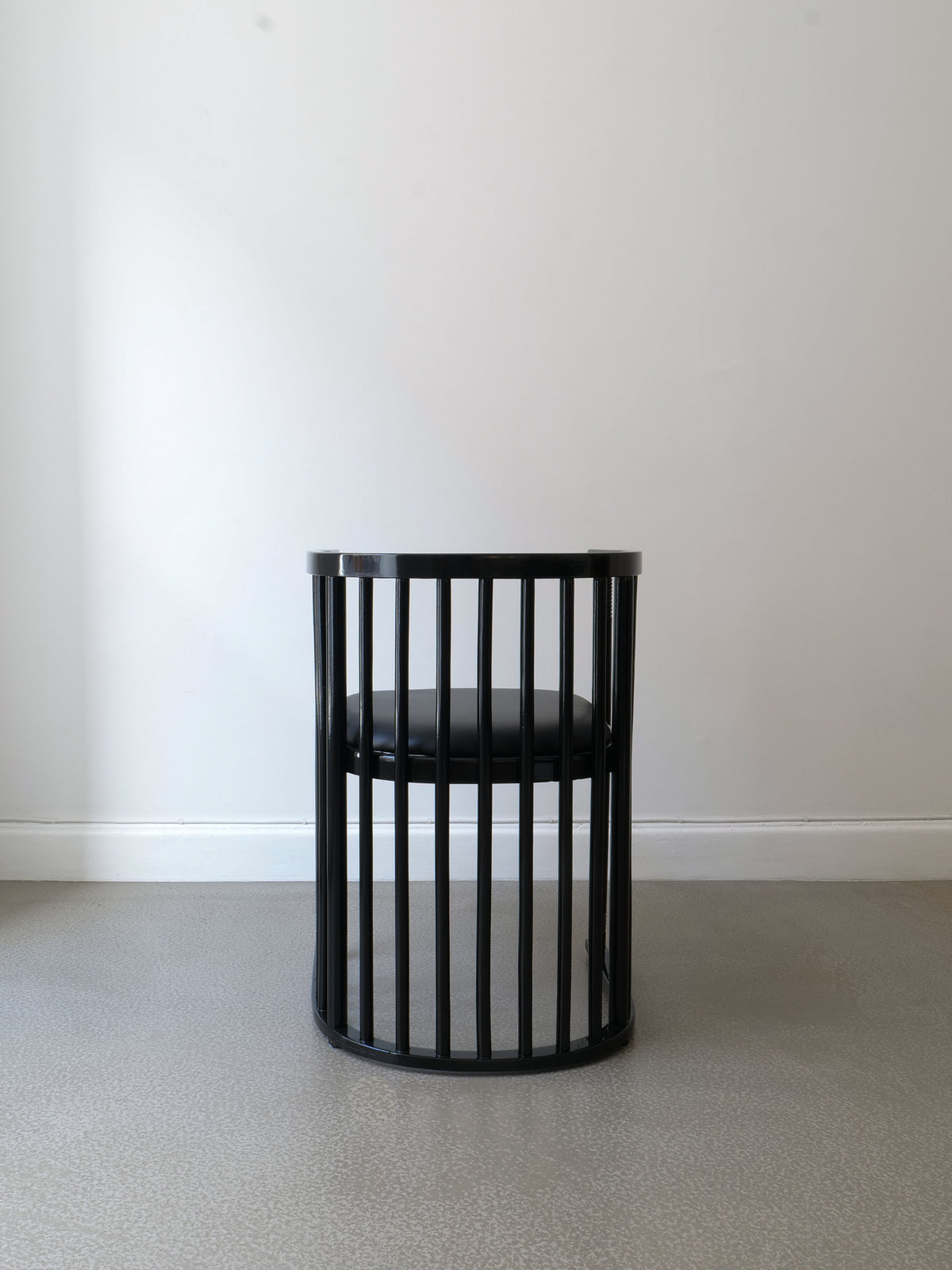 The Bentwood Armchair Josef Hoffman by Collection apart, a black, circular chair influenced by Josef Hoffman's designs, showcases tall vertical bars for a backrest and a plush padded seat. It stands elegantly on a smooth gray floor against a plain white wall.