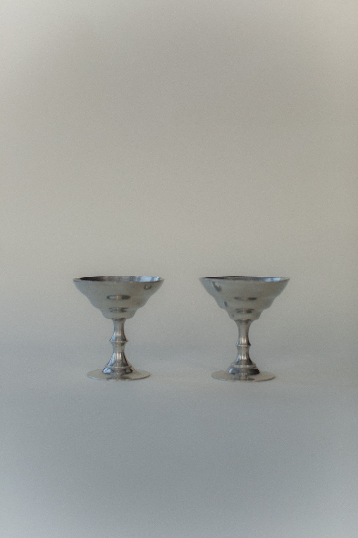 Two silver vintage-style ice cream cups from Out For Lunch, with scalloped edges, stand side by side on a smooth light gray surface. Reminiscent of Art Deco style, the softly blurred background enhances the minimalist elegance of the scene.