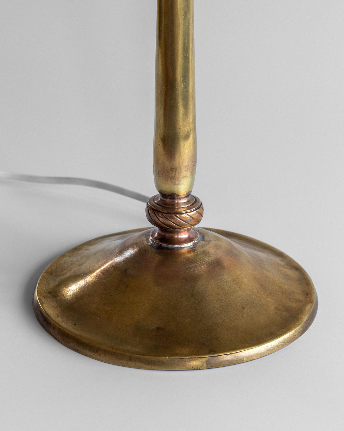 Close-up of Spigel's Art Nouveau Brass Table Lamp, showcasing a wide round base with floral engravings and a slender stem. A cord trails elegantly from the base against a light gray backdrop.