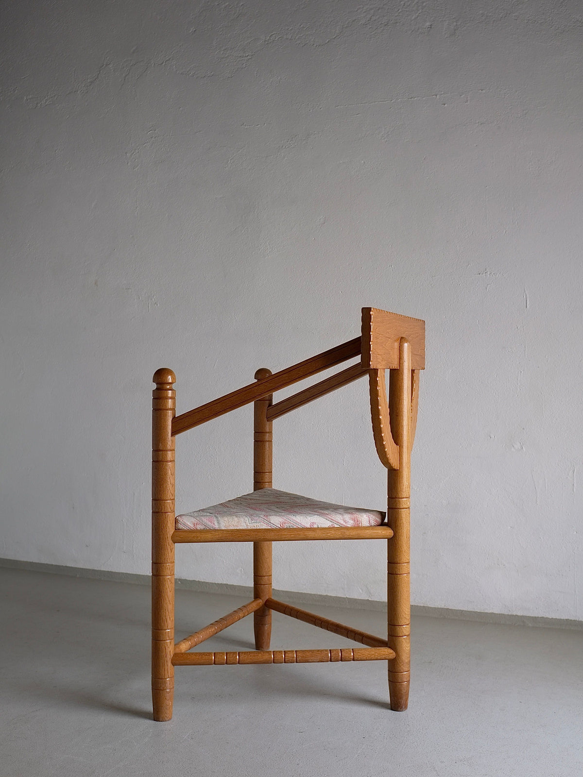 The image displays the "Rustic Carved Oak Corner Chair" from the 1950s Swedish Design period, available from Veter Vintage. The chair features cylindrical legs, a triangular base, and a horizontally flat rectangular backrest. The cushioned seat is upholstered in a light-colored fabric adorned with a subtle floral pattern. The backdrop consists of a plain white wall.