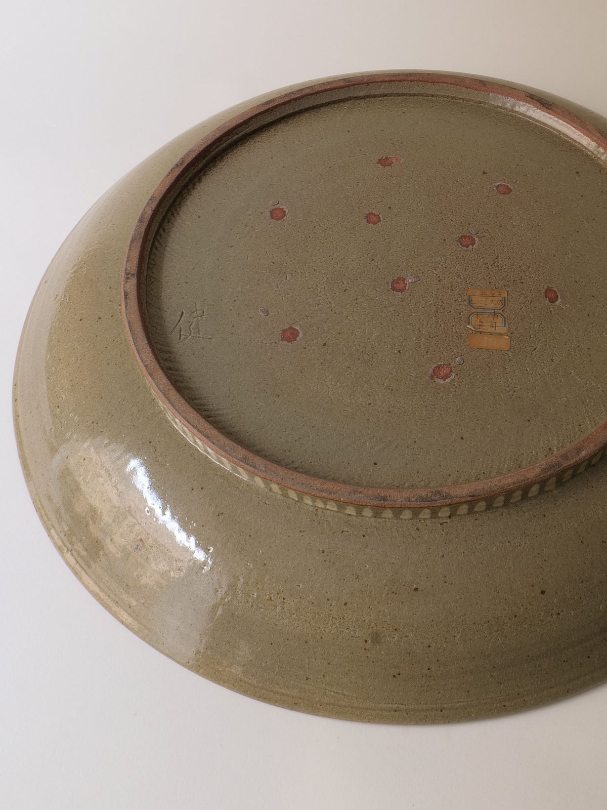 The image depicts the underside of a Ceramic Plate by Kenji Sugiura from 1980s Japan, offered by Collection apart. It has a greenish glaze akin to Shōwa-era ceramics, with small red dots at its center, rectangular labels near its edge, and a textured surface.