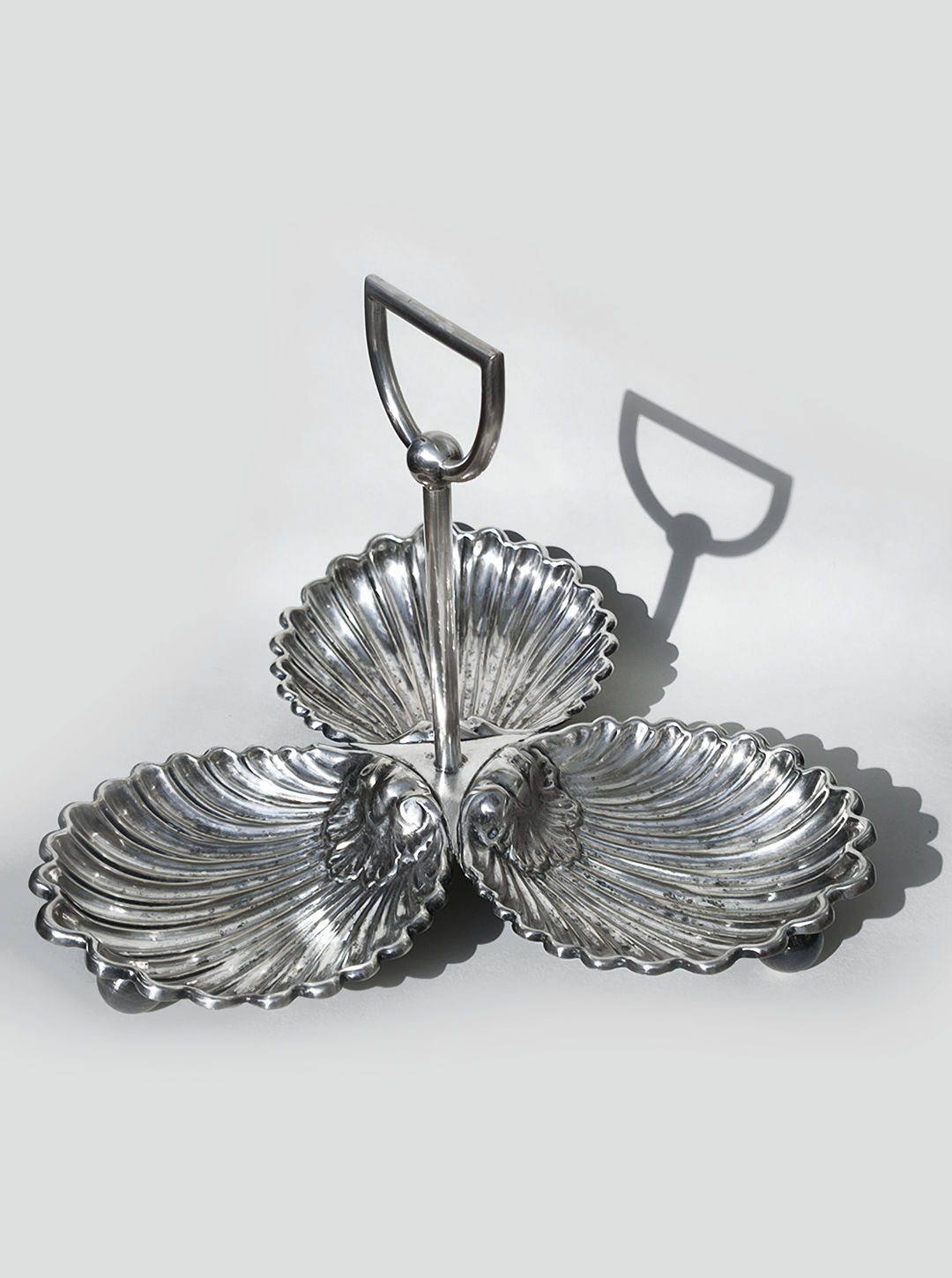 The Les Objoies Large Silver-plated Triple Tray features shell-shaped compartments and a central handle, casting a distinct shadow on a light gray background.