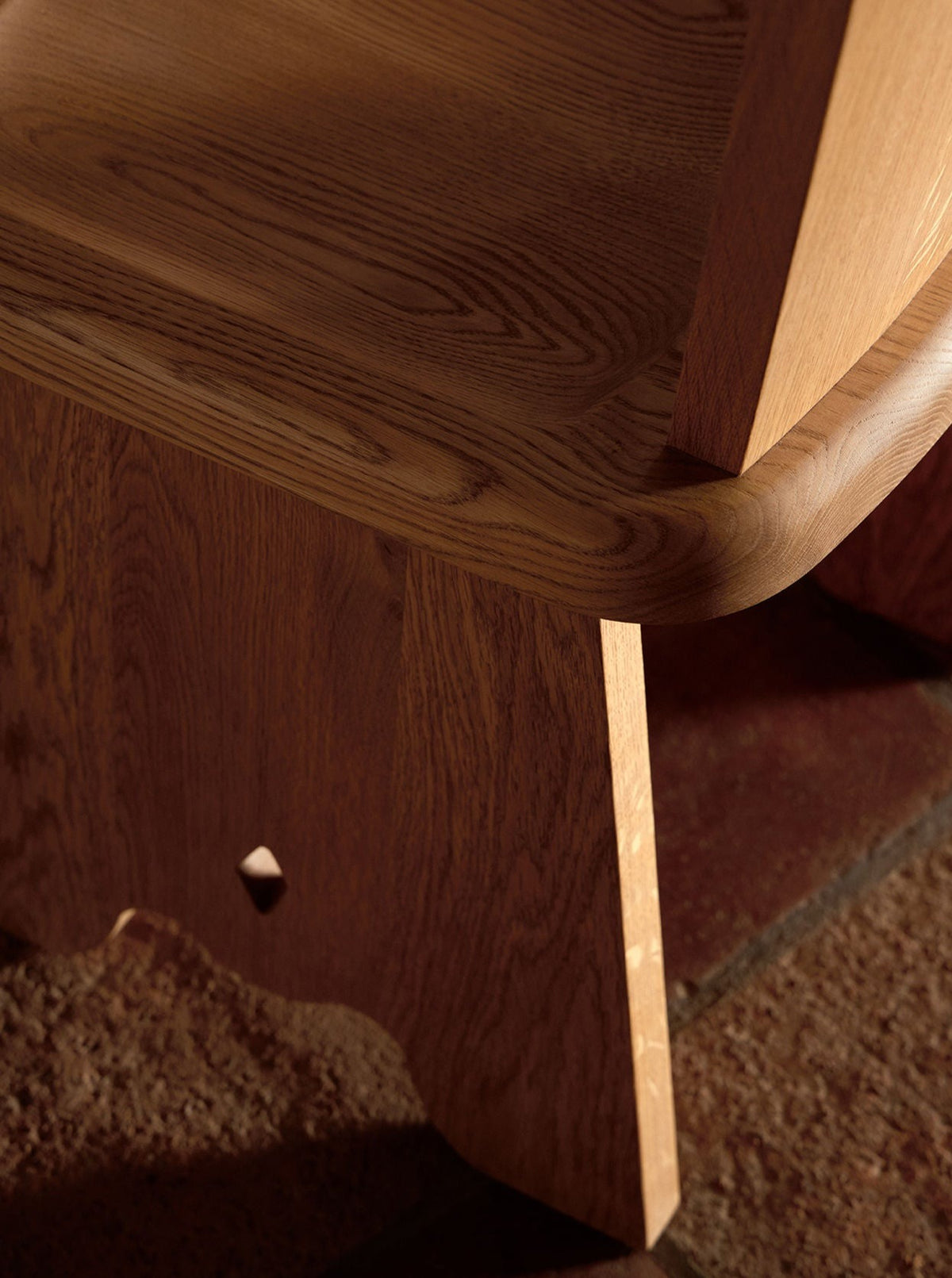 Close-up image showing the corner of a wooden chair with intricate grain patterns, highlighting the smooth finish and craftsmanship. The Rambling Chair in Natural French Oak Wood by Lemon is placed on a textured carpet, contributing to the overall warm and cozy ambiance.