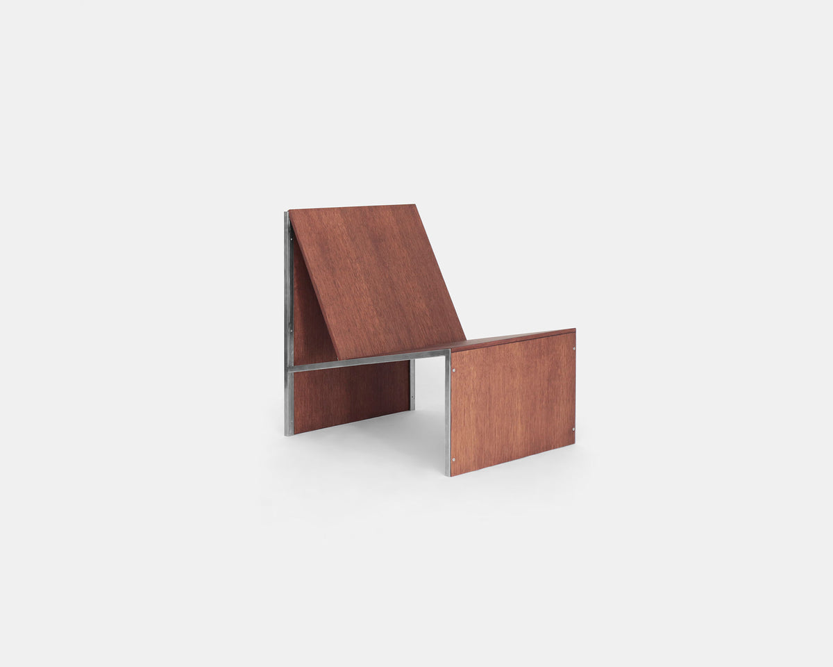 The Lounge Chair 2022 by Studio HAOS features a minimalist wooden design with slanted backrest and seat, supported by a simple metal frame, showcasing grounded elegance against a plain light gray background.