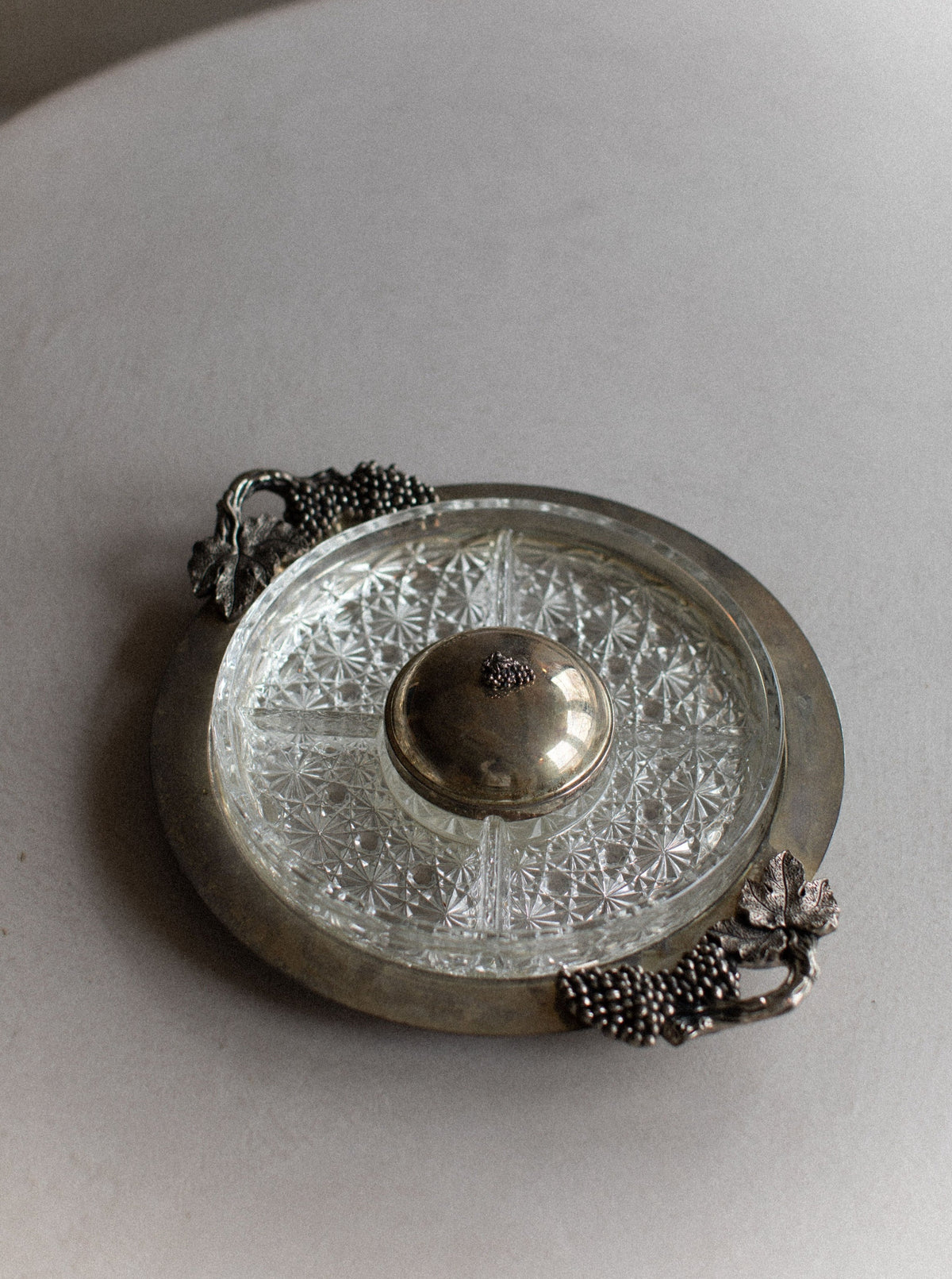 An Art Deco Tray-set by Out For Lunch with a silver-colored metal rim and two ornate grapevine handles. The steel and glass base features an intricate geometric pattern, and a small metal dome lid rests at the center, epitomizing timeless elegance. The tray sits on a smooth, light gray surface.