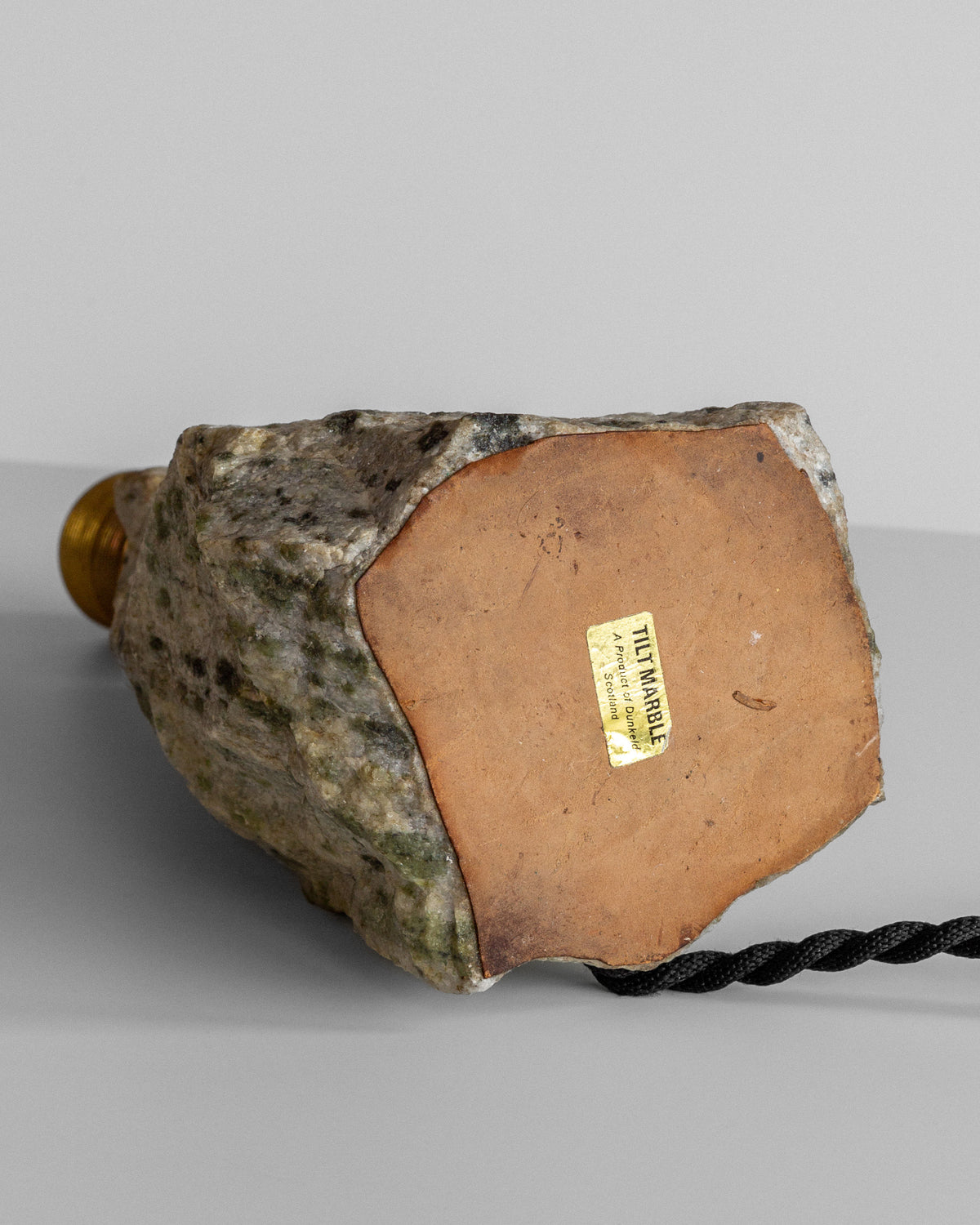 The **Spigel Marble Stone Table Lamp** with a rough stone texture and an obelisk shape sits on a surface. A flat base features a gold rectangle sticker with text, and from the base extends a black, twisted electrical cord. The background is plain light gray.