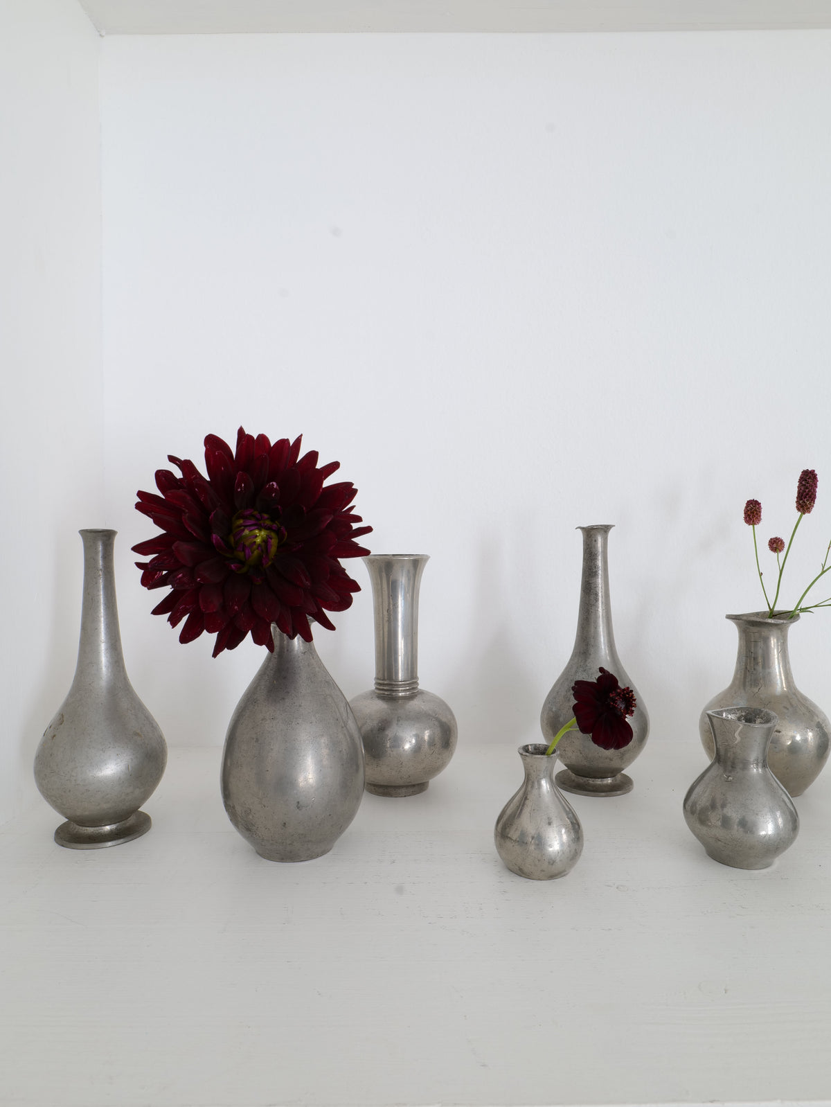 The Just Andersen Mini Vase Collection by Collection apart, inspired by Danish silversmith designs, is elegantly showcased on a white surface against a white background. Some vases hold deep red flowers, infusing vibrant contrast into the minimalist display and adding an extra touch of charm.