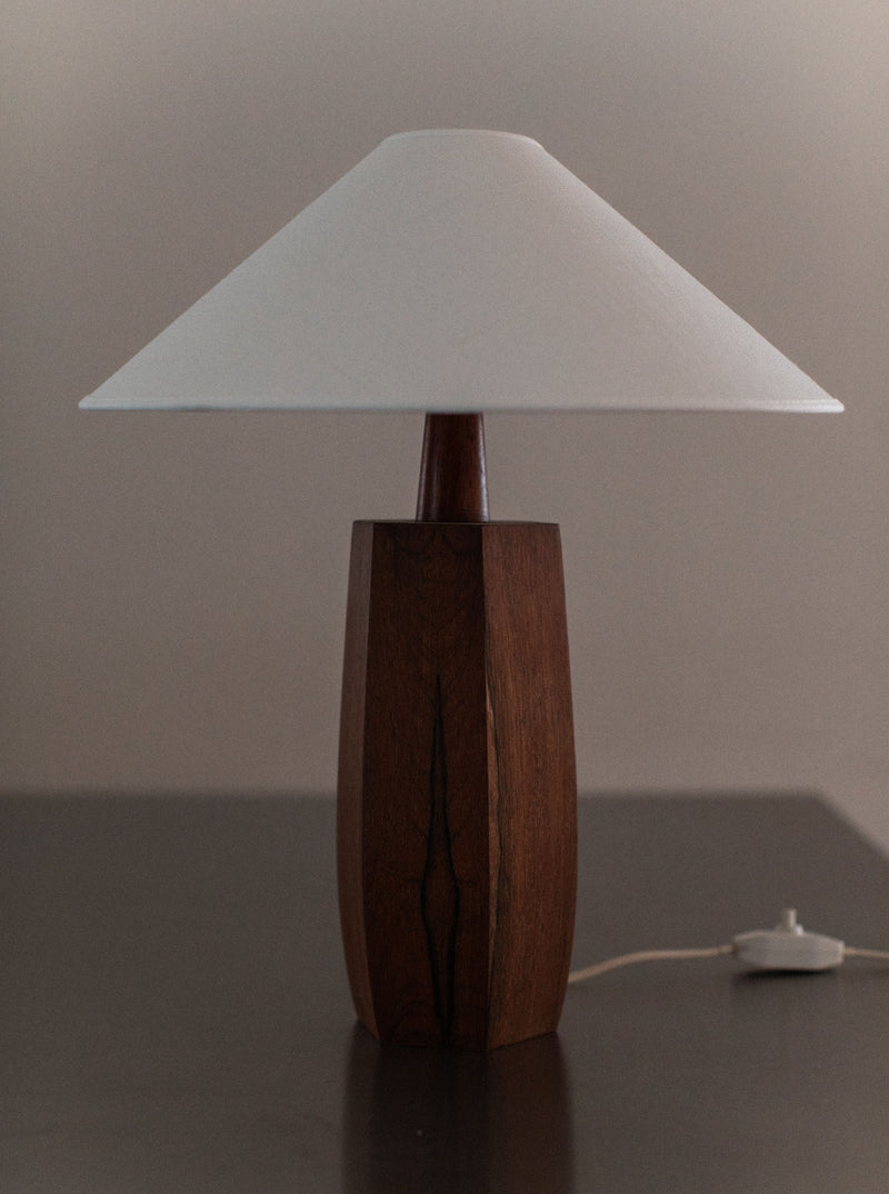 A 1940's Wooden lamp from Out For Lunch, showcasing mid-century design with a white, conical shade, stands on a dark surface. The lamp's cord and switch are visible at the base, extending towards the right. The background is neutral, highlighting the lamp's simple and elegant craftsmanship.