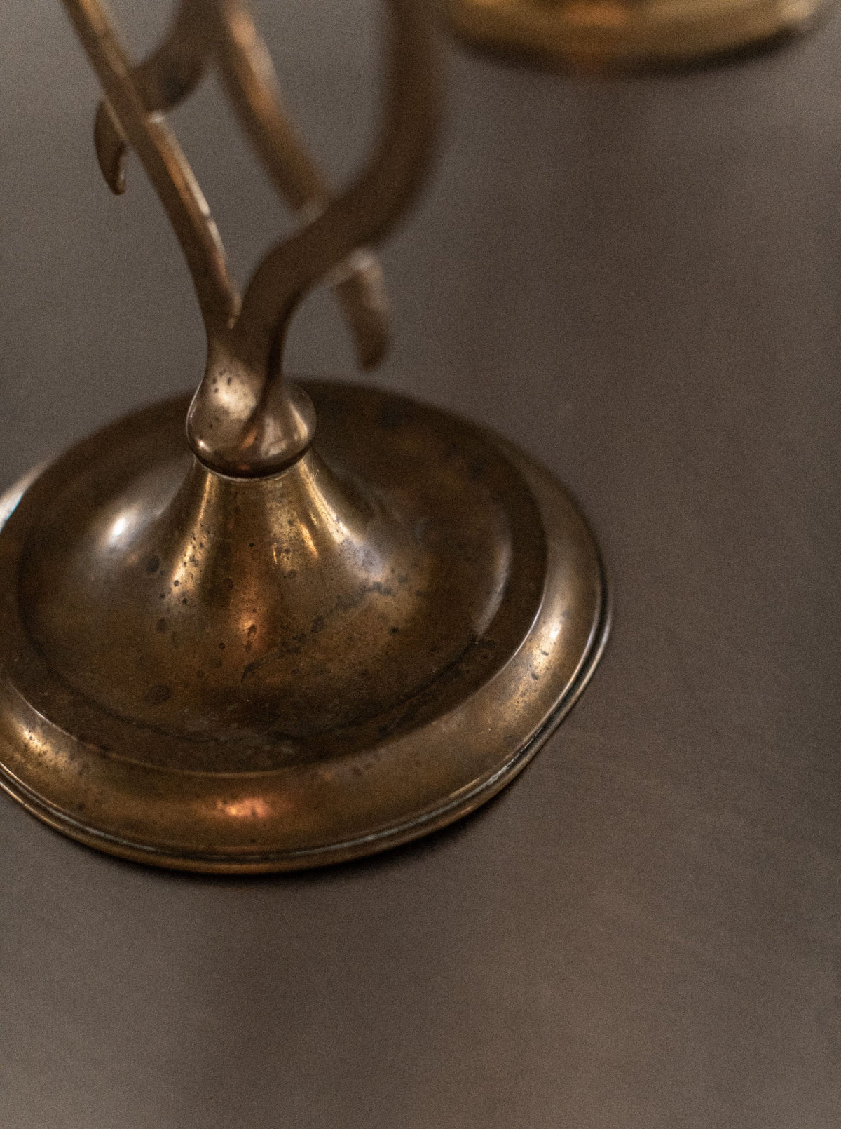 A close-up of the base of the Out For Lunch Brass Candleholder resting on a smooth, reflective surface reveals its aged character and usage marks, highlighting its unique history. The patina on the metal showcases its antique charm. The elegant curves of this timeless piece make it a classic addition to any home decor.