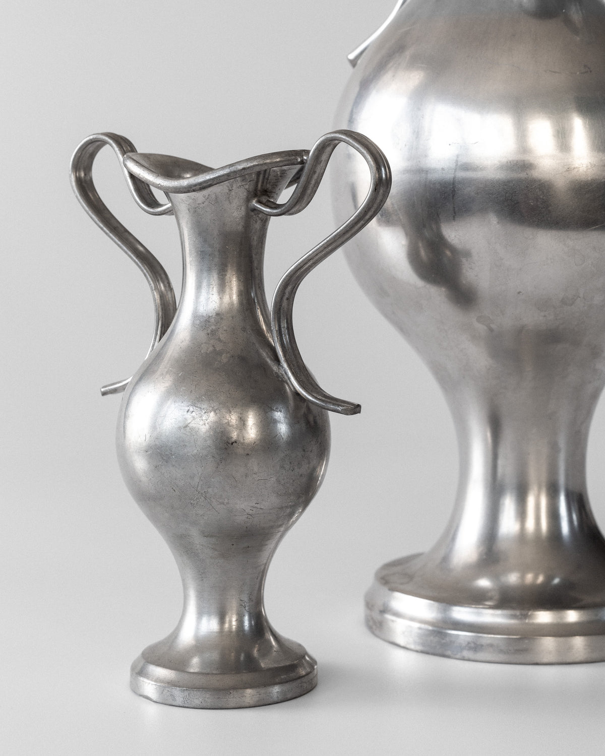 A close-up features two Spigel neoclassical metal vases from 1953, Sweden, with ornate handles. The polished silver finish reflects light beautifully against a plain white backdrop.