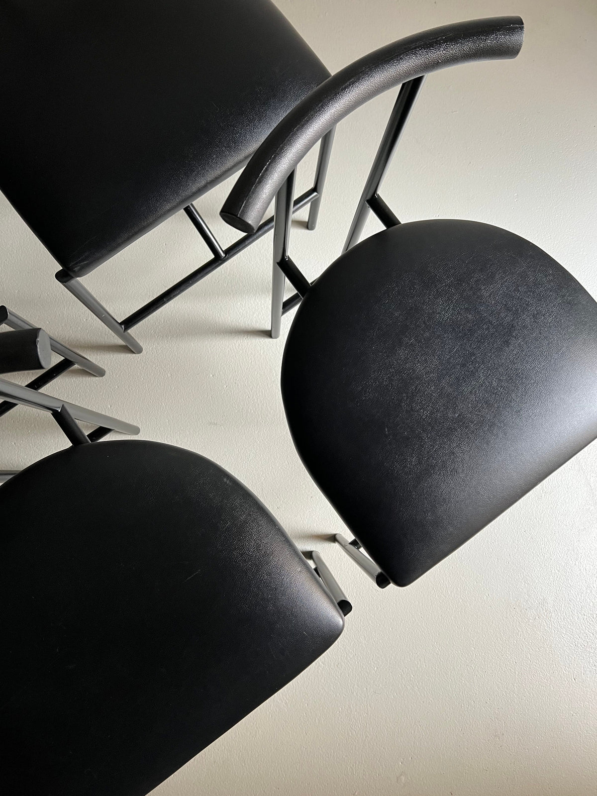 A view from above four 4 Postmodern "Tokyo" Chairs by Rodney Kinsman | Italy | 1980s arranged in a seemingly random manner. The seats are curved, featuring faux leather upholstery that lends them a sleek yet vintage condition. The chairs' legs are thin and angular, casting subtle shadows on the light-colored floor.