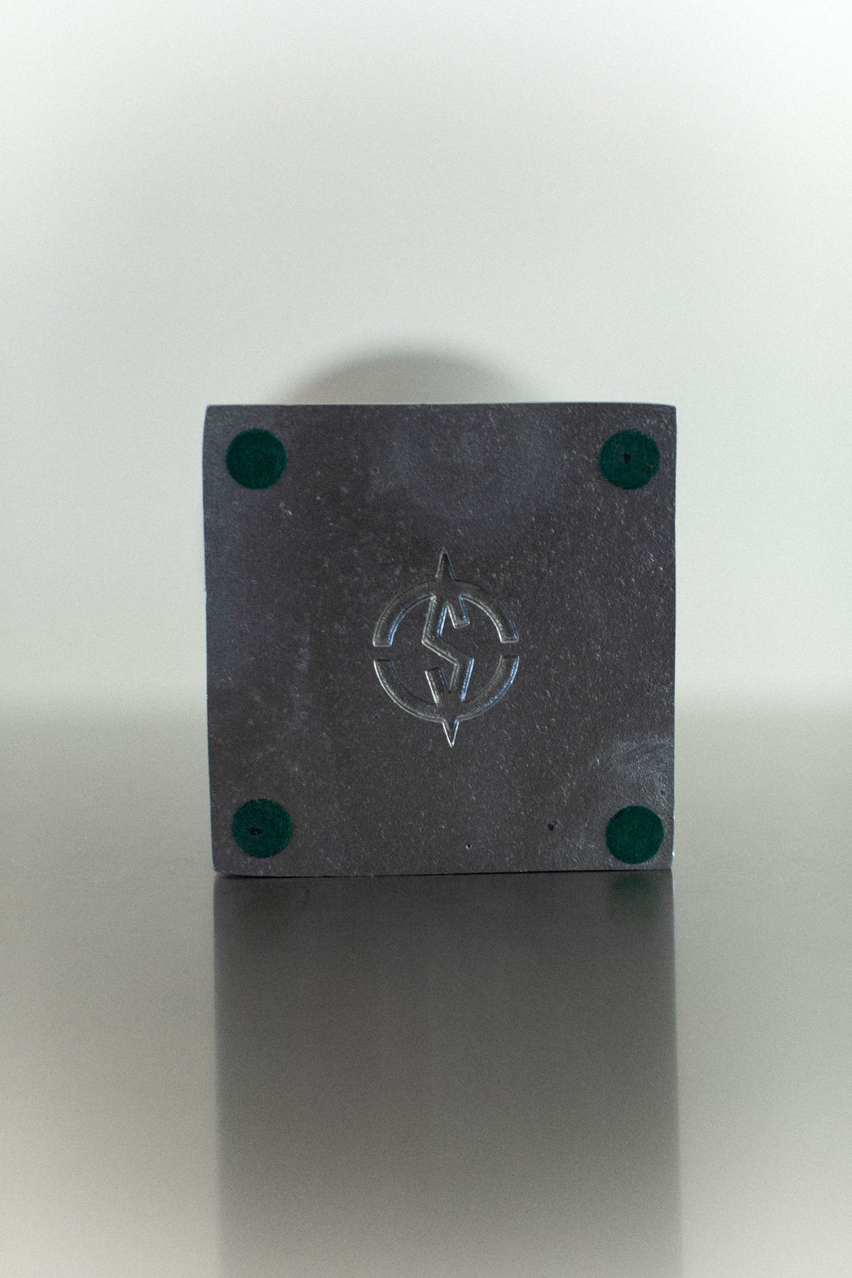 A square, gray coaster adorned with a lightning bolt and circle logo conveys a modern design, highlighted by its four green circular pads at each corner. It rests on a sleek surface, reminiscent of the 70's Aluminum Cast Vase from Out For Lunch.