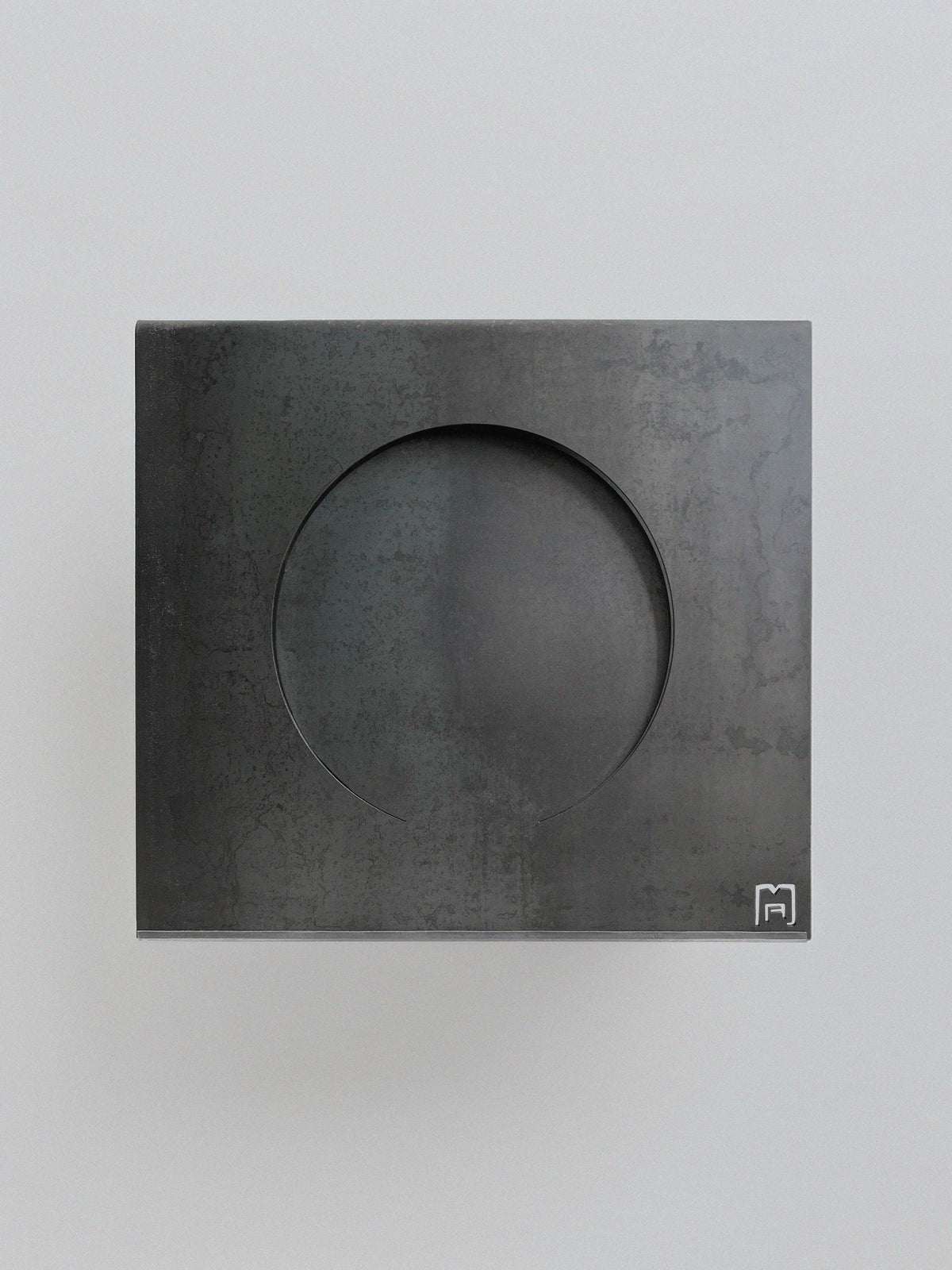 The 'Era' Side Table by Modan features a square steel surface with a large circular indentation in the center and a small, white-outlined icon at the lower right corner, set on a light gray background for a distinctive look.