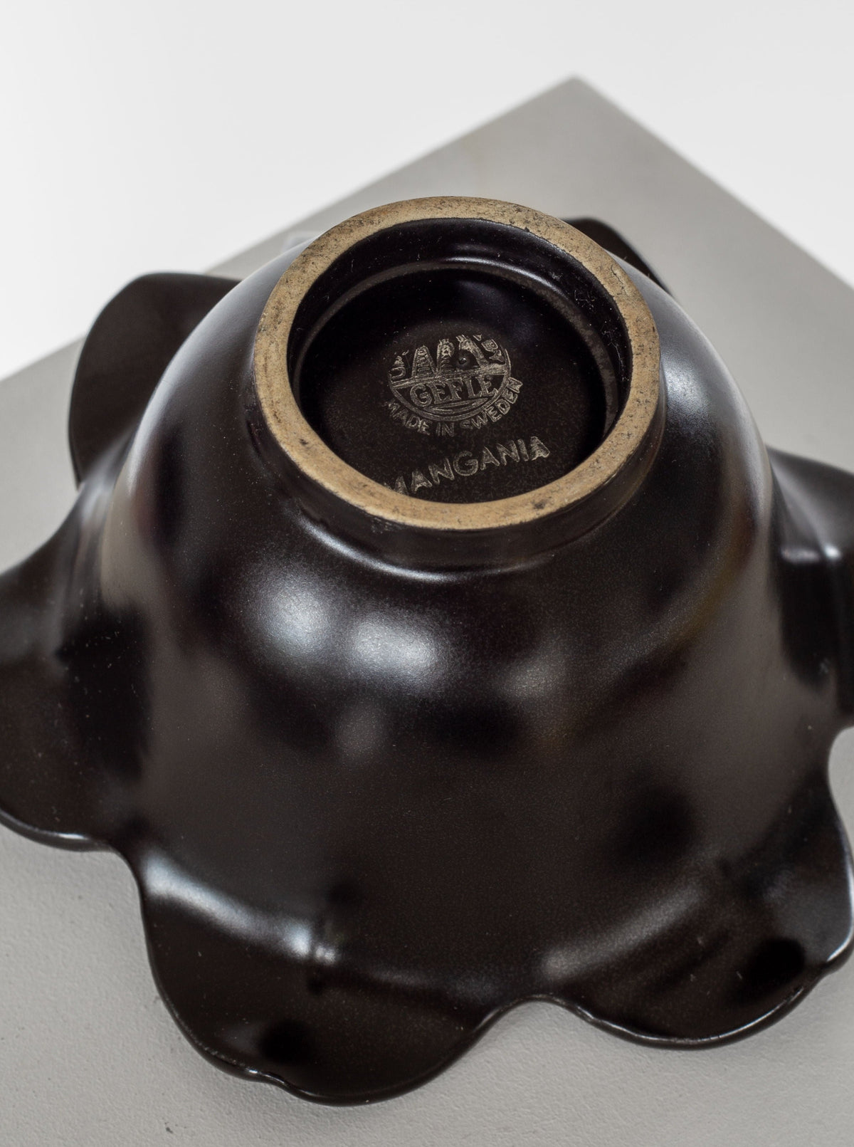 A close-up shot of a dark, glossy bowl upside down, placed on a table. The bowl's bottom is marked with "MADE IN ITALY" and "MANGANIA" along with an embossed emblem. Reminiscent of Swedish Mid-Century Modern studio pottery—much like the Spigel Mid-Century Ceramic Candlesticks from the 1950s—the bowl's edges have a wave-like pattern.