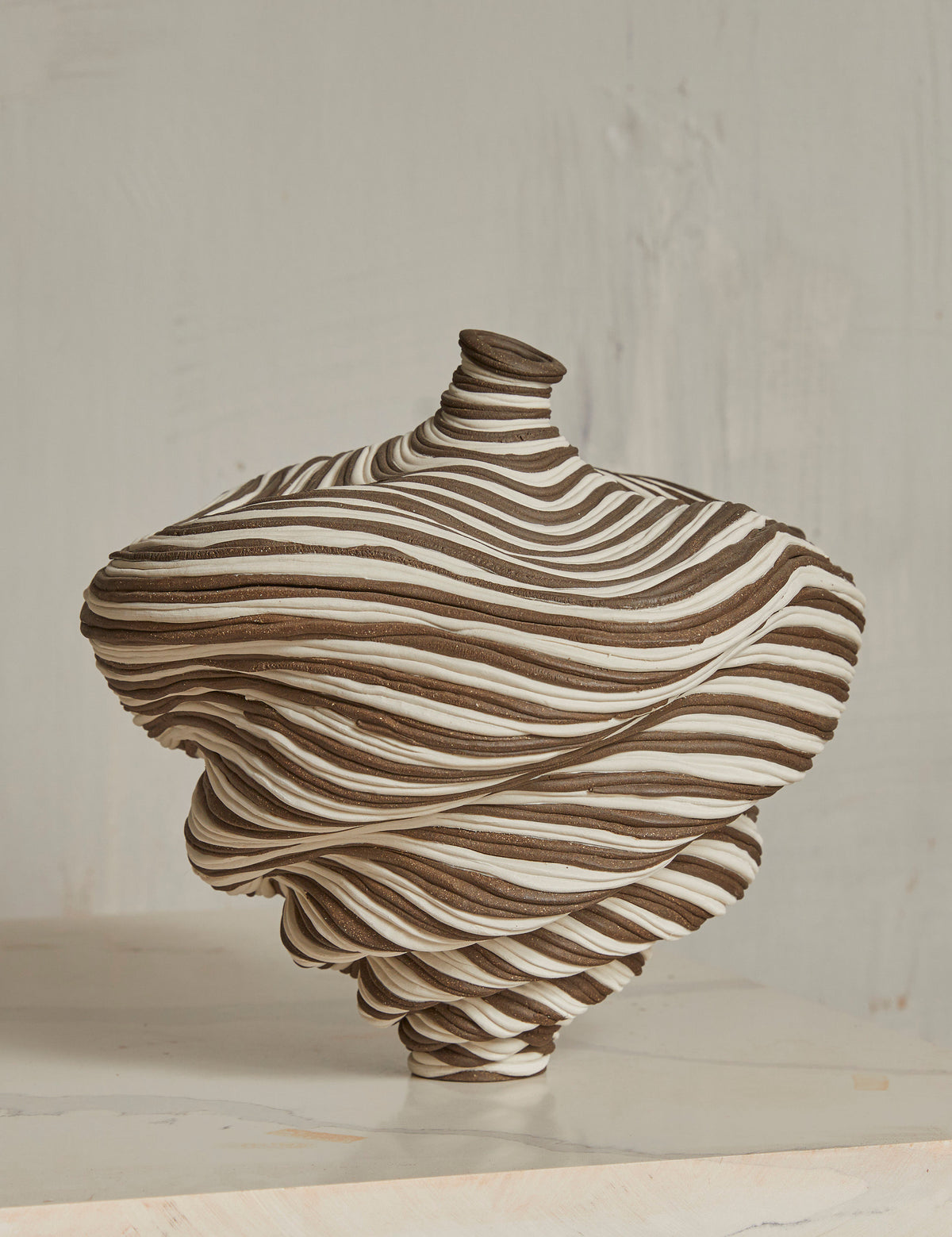 The Soucy Shop's Black & White Bottle is a ceramic vase crafted with stoneware coils, showcasing a twisted pattern of dark brown and white stripes. It resembles a distorted spiral and rests on a light marble surface against a muted, textured wall.