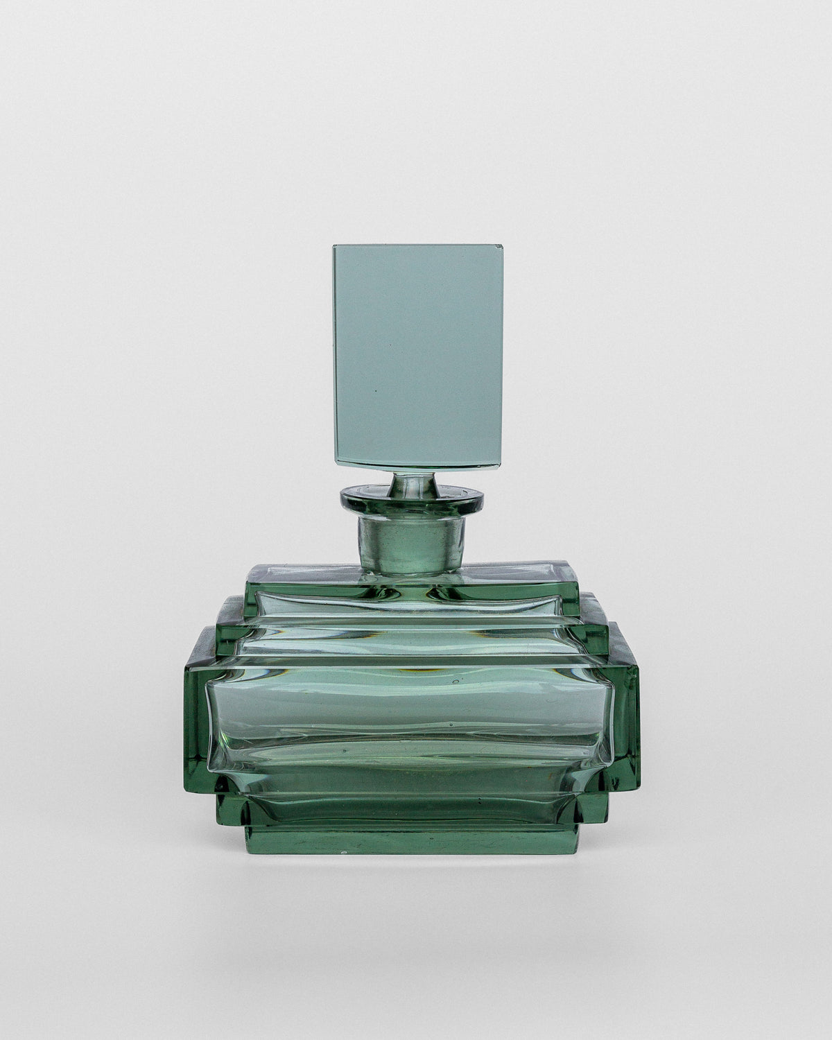 The Spigel Art Deco Perfume Bottle, 1930s, is a rectangular, transparent green piece with a matching green cap. It boasts a geometric design inspired by the aesthetics of the 1930s, featuring clean lines and sharp edges for a modern and elegant appearance. The style suggests possible Czech origins against the plain white background.
