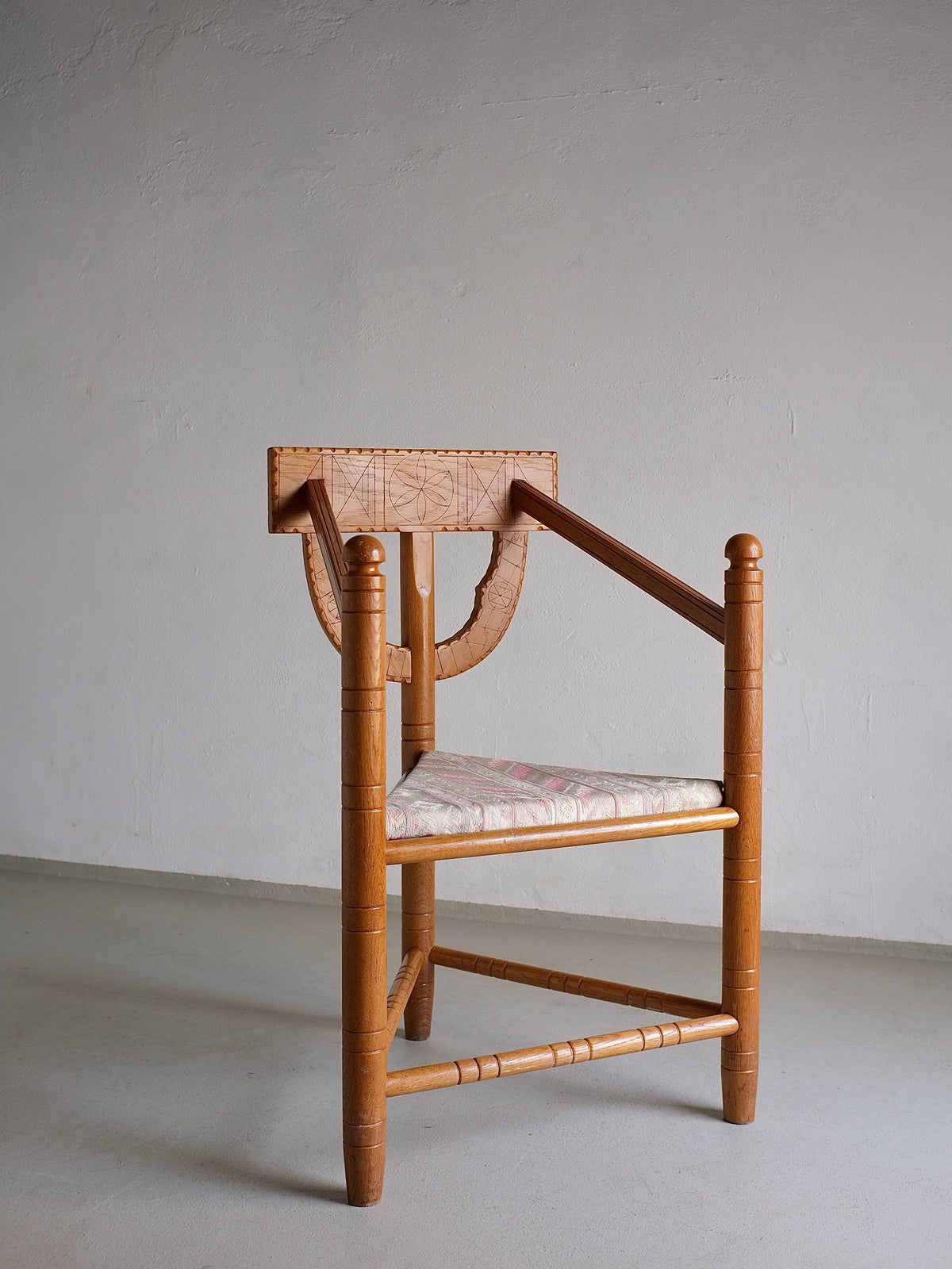 The Rustic Carved Oak Corner Chair from Veter Vintage features a unique, vintage design with a geometric backrest and angled armrests. This wooden chair has a patterned cushioned seat and spherical accents on the legs, reminiscent of Sweden's 1950s design period. It is pictured standing against an off-white wall with a gray floor.