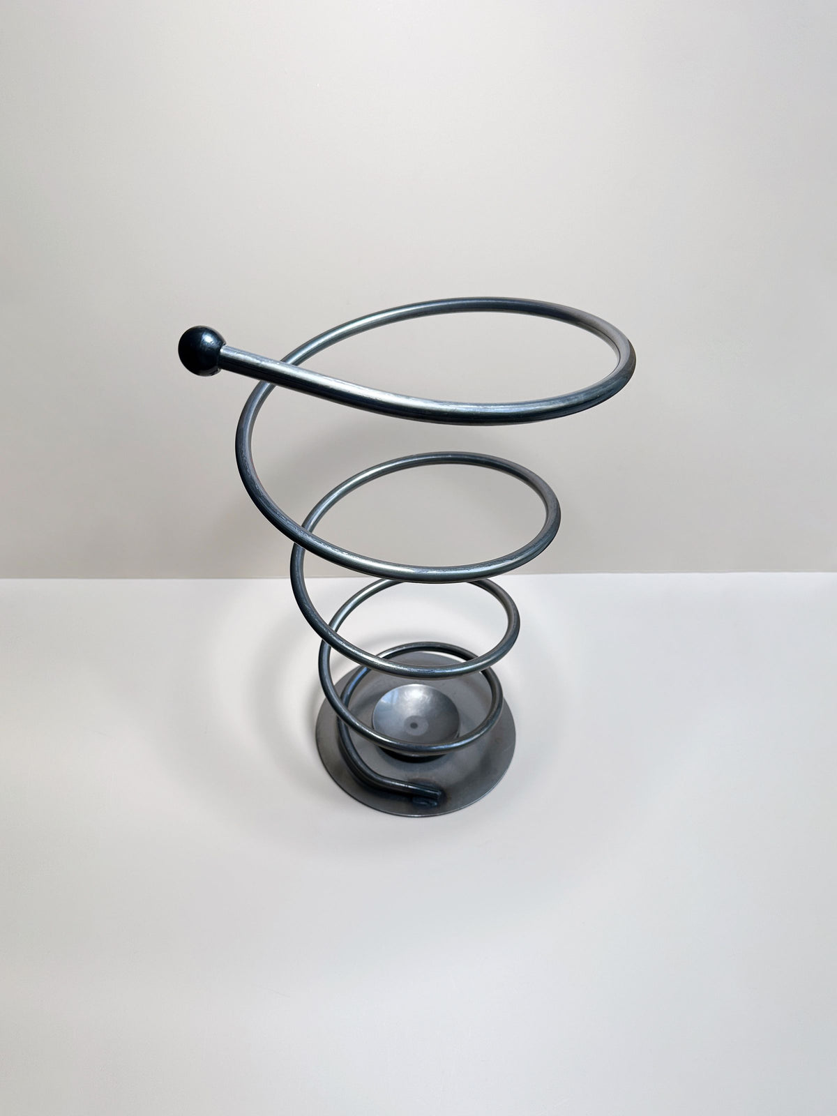 The Dodo Vintage Spiral Metal Umbrella Stand, with its twisted, spiral-shaped structure and circular base reminiscent of a coiled spring, sits elegantly on a flat surface against a plain, light-colored background. This solid metal stand features an elongated loop at the top and exudes a minimalist design that evokes the aesthetic of post-modern decor.