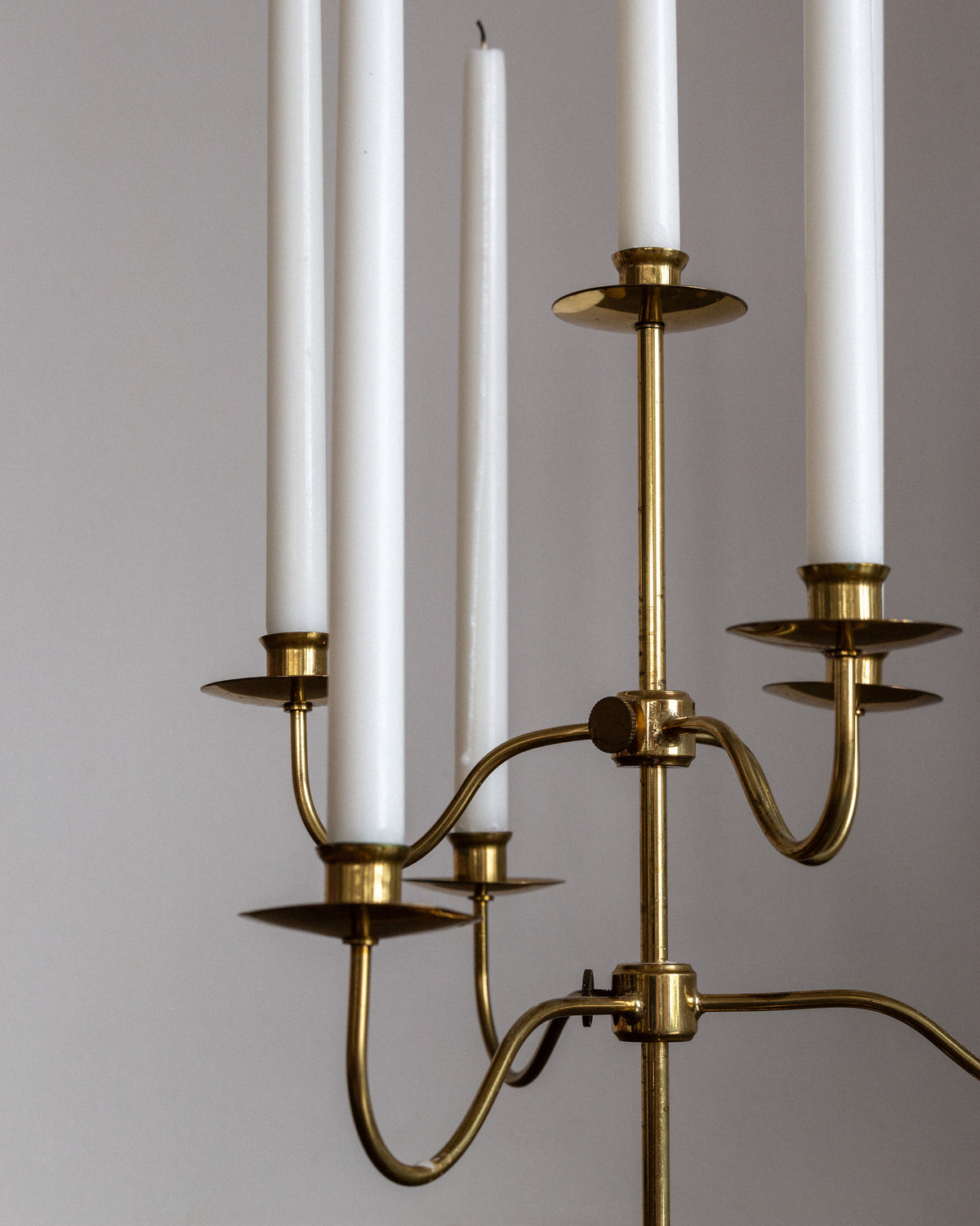A close-up displays the Spigel Swedish Mid-Century Brass Candelabra with five unlit white candles set against a plain, light gray backdrop. This piece boasts an elegant Scandinavian Modern style with curved arms and a polished finish.