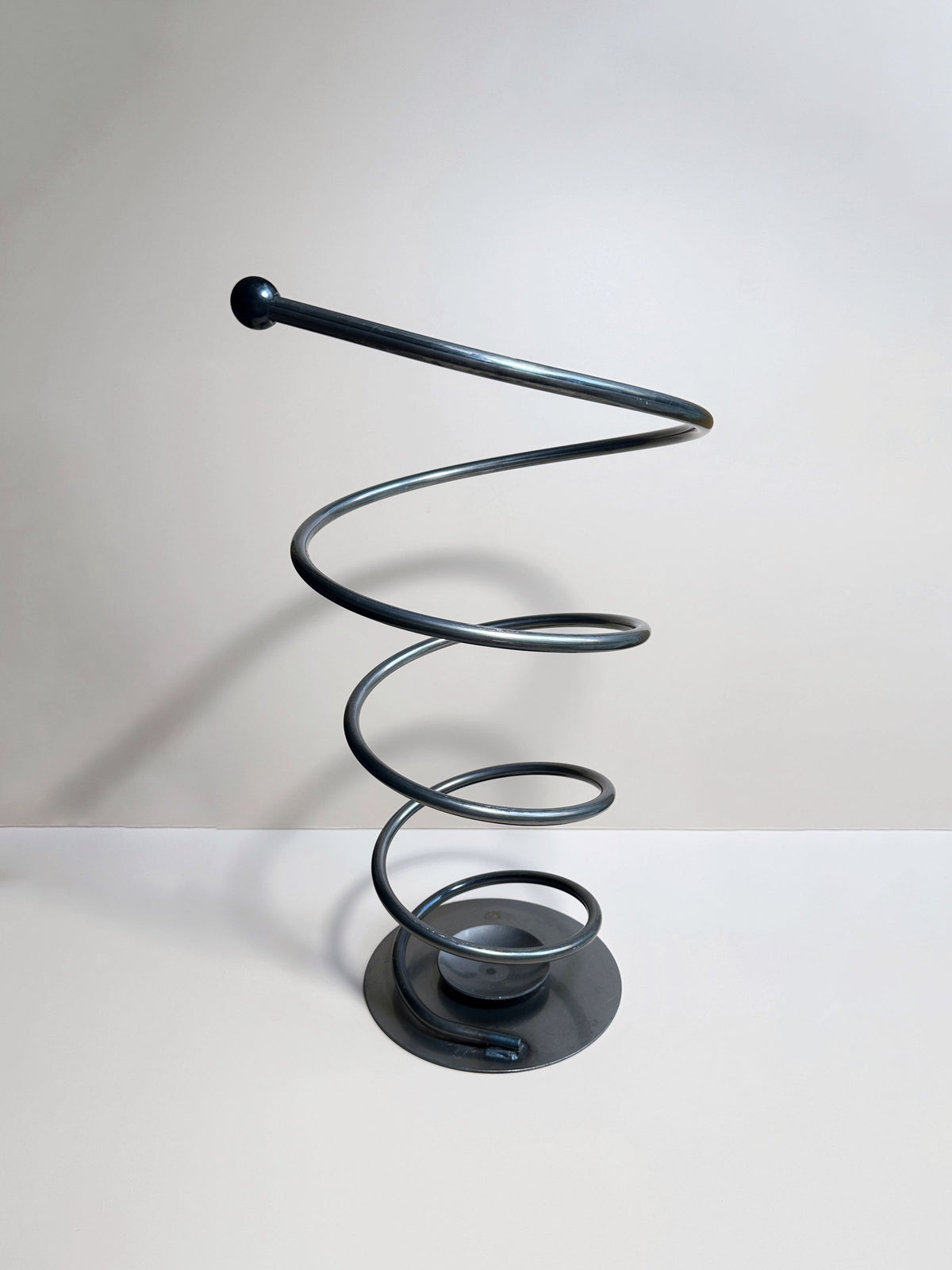 The Spiral Metal Umbrella Stand by Dodo Vintage is a modern, minimalist piece featuring a sleek black metal spiral shape extending upward from a circular base. The solid metal stand has a smooth, continuous coil with a spherical tip at the top and is set against a plain, grayish background.