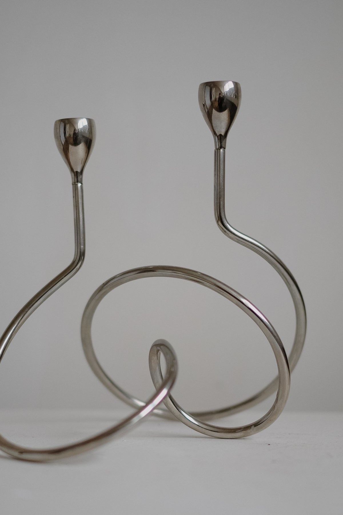 Against a plain gray backdrop, two "Loop" candle holders by septembre studios feature modern, minimalist chrome designs with sleek curves. Their unique shapes provide a contemporary artistic flair, ideal for displaying elegant dinner candles.