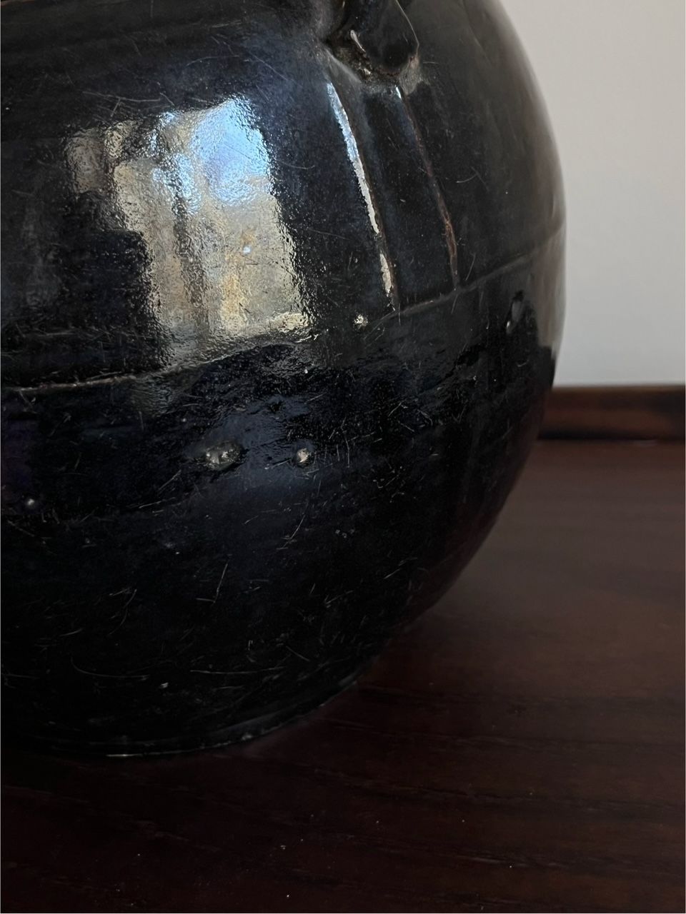 A close-up of the Antique Glazed Lamp with Ears by Médecine, showcasing its shiny black ceramic finish and rounded shape like an antique vessel, placed on a dark wooden surface. Reflective highlights reveal its smooth, polished texture.