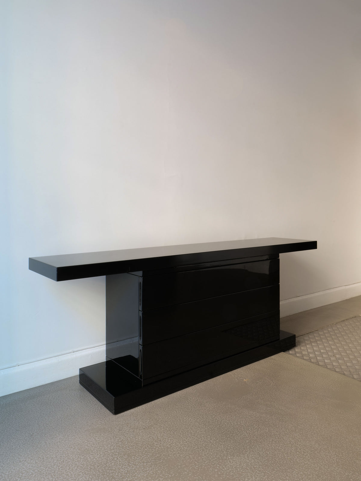 The High Gloss Console Table 1970s by Collection apart, with its modern and sleek Italian design, stands elegantly against a plain white wall. This custom-built piece showcases a minimalist style, featuring a rectangular top and sturdy base, while the floor highlights a light, textured surface.