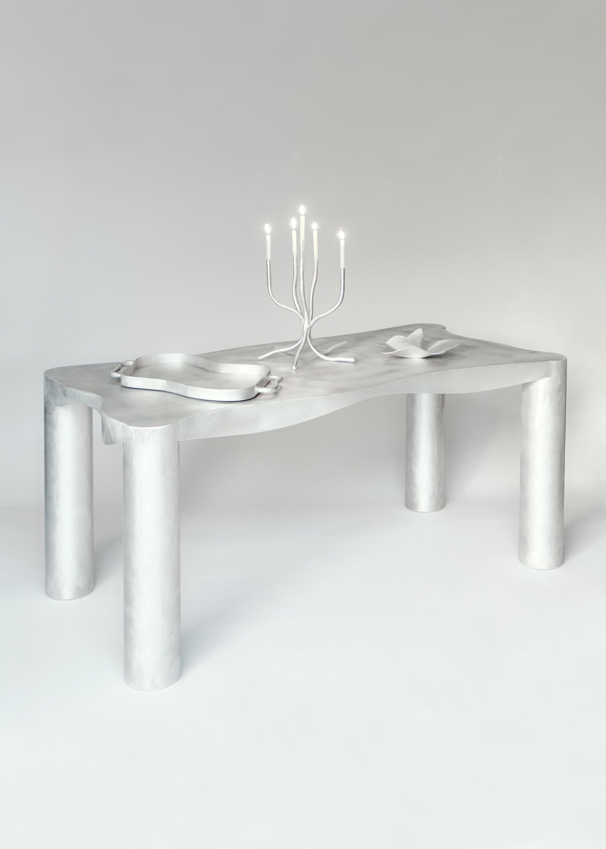 Crafted in a North London workshop, this minimalist white serving tray by Six Dots Design features wavy edges and thick cylindrical legs. The elegant, monochromatic scene is enhanced by two stylish plates with ribbon-like designs and a modern candelabra with lit candles, making it an exquisite choice for any setting.
