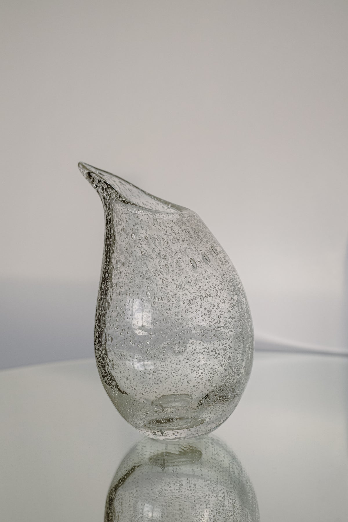 The Small Bubble Vase by septembre studios features exquisite craftsmanship with a curved, textured, clear glass design and a unique organic shape, beautifully displayed on a reflective surface against a minimalist background.