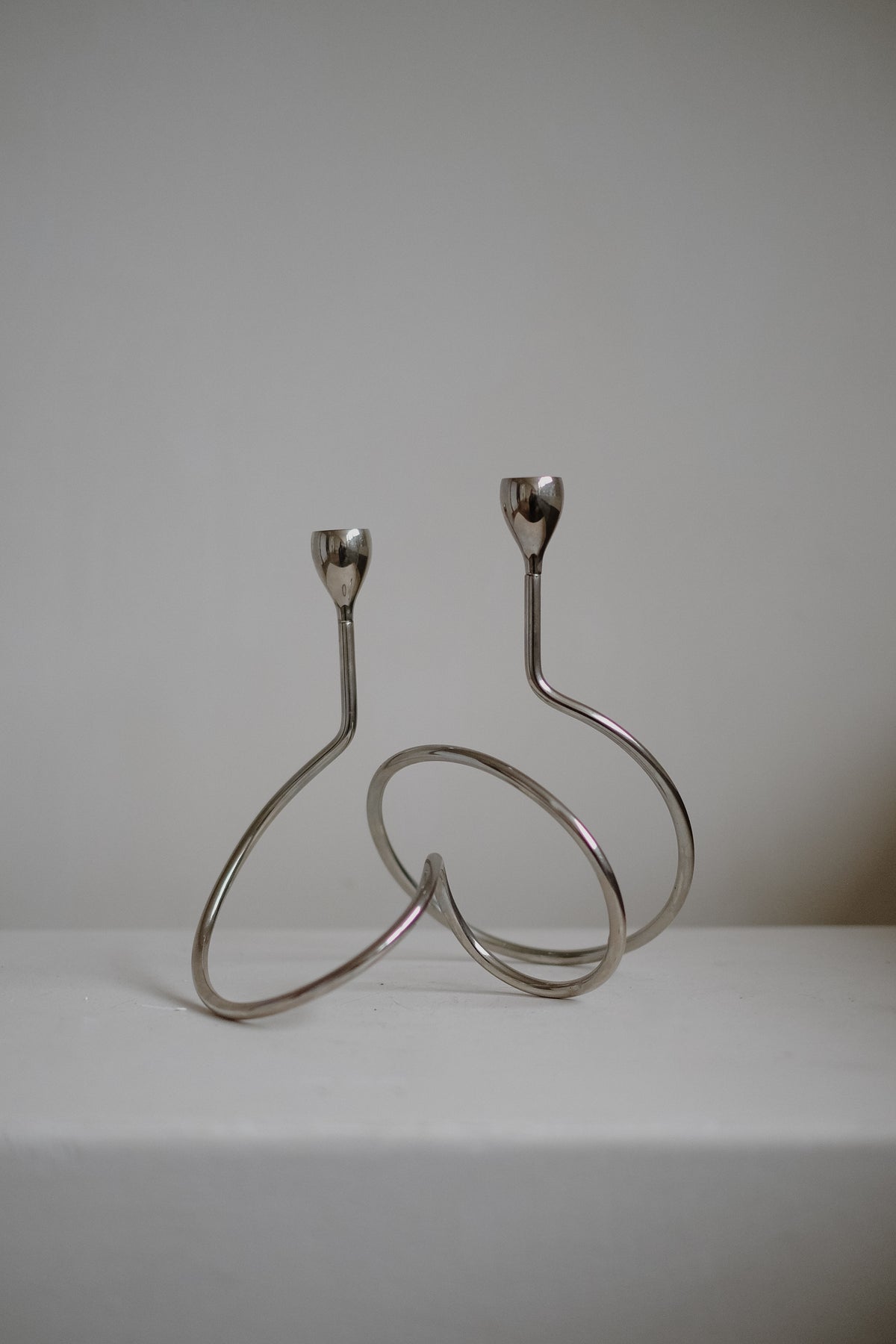 A pair of "Loop" Candle Holders by septembre studios, with a modern chrome, twisted abstract design, sit on a white surface against a plain backdrop. Each has a minimalistic, curvy shape perfect for elegant dinner candles.