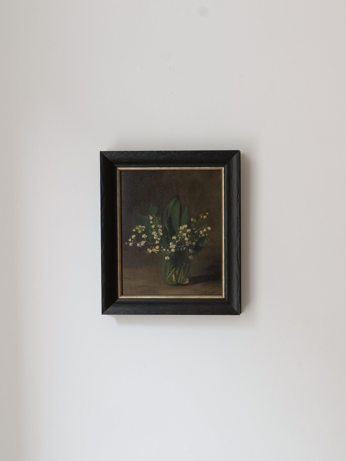 A framed Lily of the Valley Still Life 1961 painting by Collection apart hangs on a plain white wall. The dark frame contrasts with the light background, highlighting the delicate white blooms and green leaves in their green vase, reminiscent of Dutch still life art.