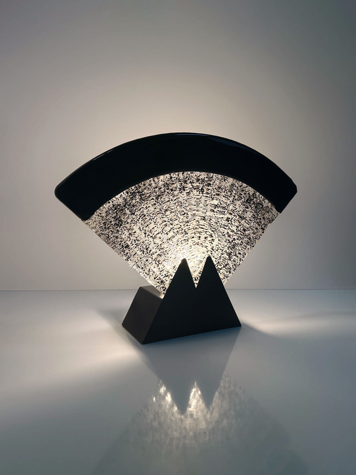The Large Murano glass fan lamp by Dodo Vintage is a 1980s Art Deco piece with a black curved top, textured translucent center, and triangular black base; it resembles a modern stylized fan and glows from within on a glossy surface.