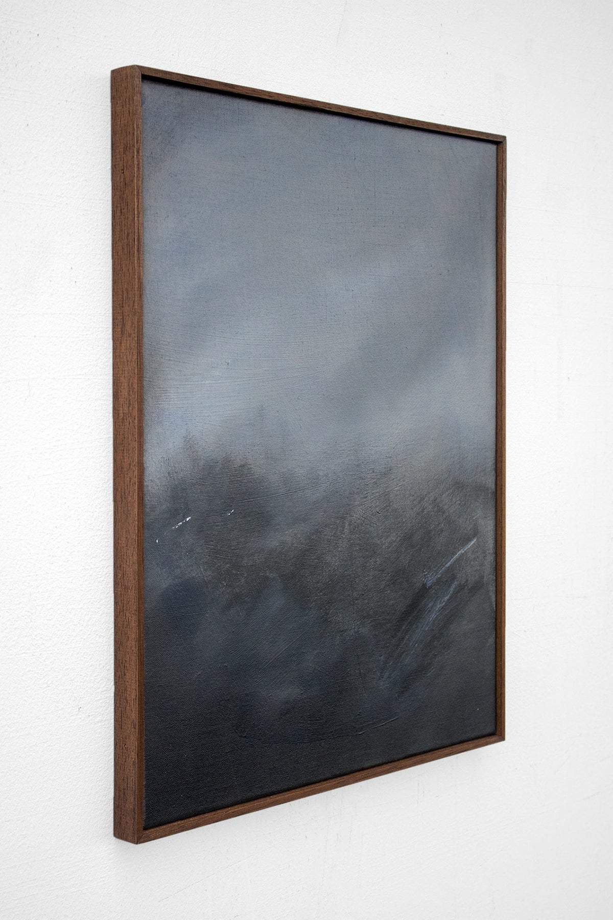 As Night Draws In by David Hardy is an original abstract acrylic on linen, featuring dark, smoky tones of black, gray, and blue that evoke a moody landscape. Framed and displayed on a white wall, it’s viewed from an angle.