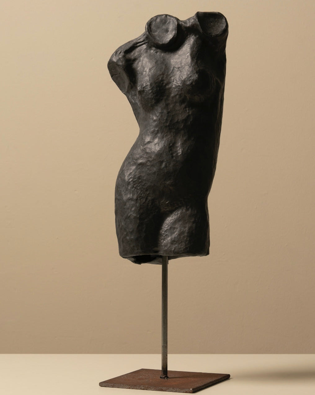 The Zinc Torso Sculpture by Les Objets Amsterdam features a dark, textured vintage female form without arms or head, mounted on a metal stand with a rectangular base, displayed against a neutral beige background.