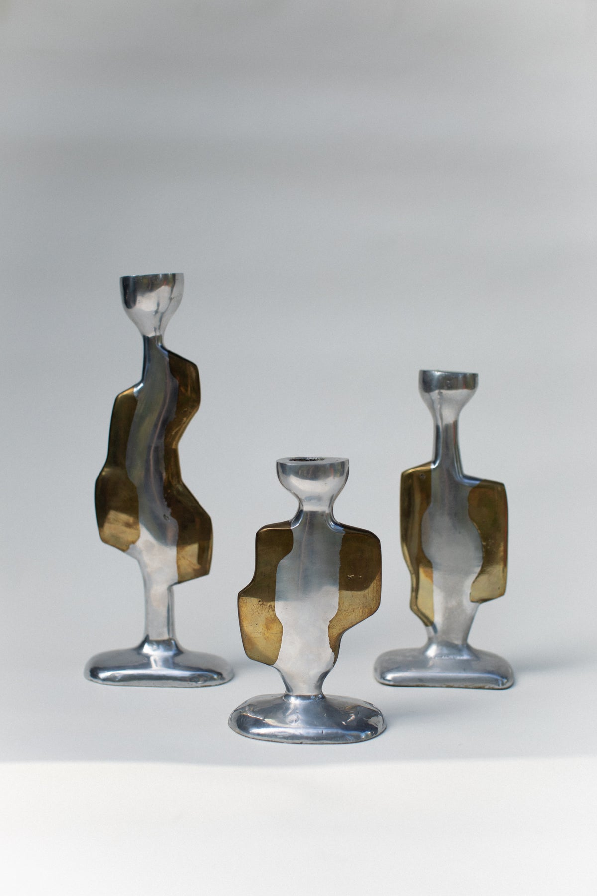 Three Brutalist Candleholders by Out For Lunch, featuring abstract wavy shapes and bronze-colored accents, are arranged on a neutral background with soft, natural lighting.