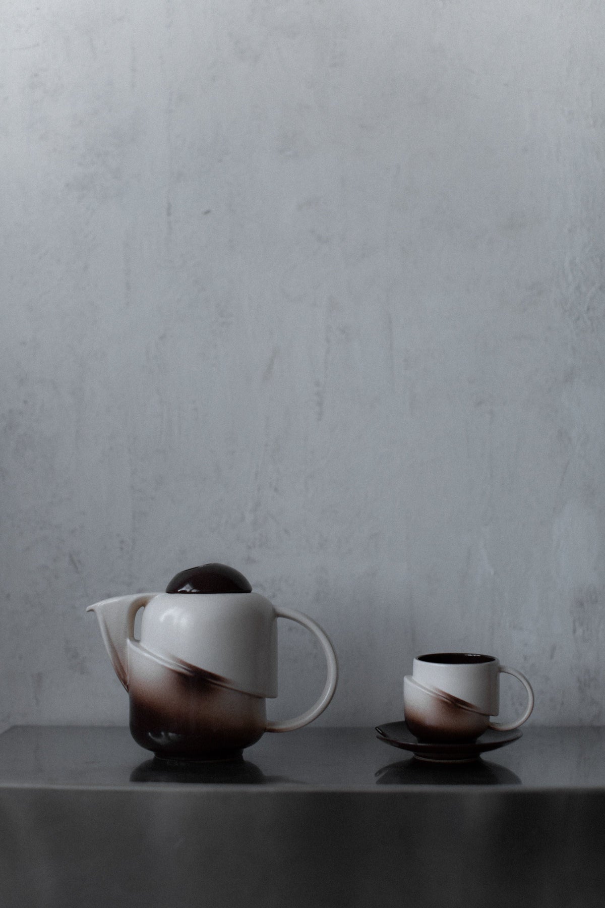 A minimalist scene with the Out For Lunch Art Deco Ceramic set—vintage teapot and tea cup with brown and white swirls—displayed on a reflective surface against a textured light grey wall.