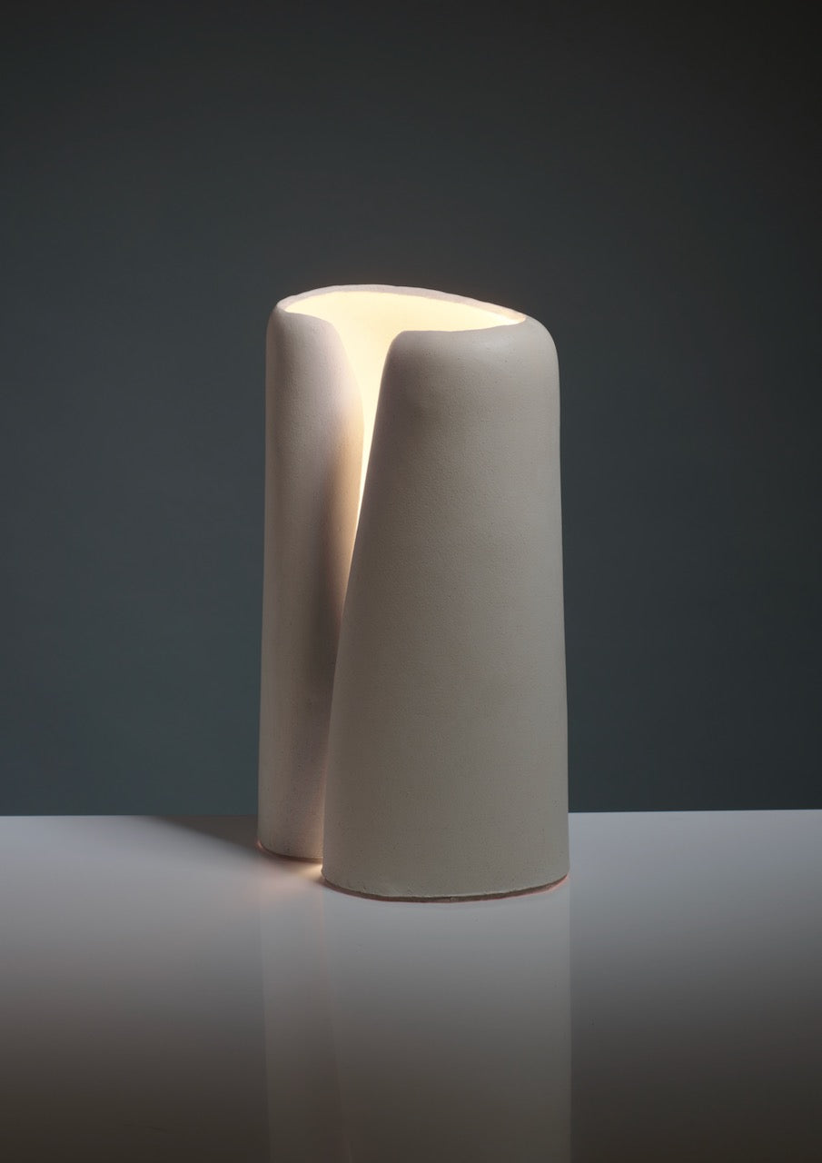 The Snail Table Lamps in Ceramic by Katia Langeard feature a modern, sculptural design with smooth matte curves that emit a warm glow—ideal as a unique bedside lamp on reflective surfaces against dark backgrounds.