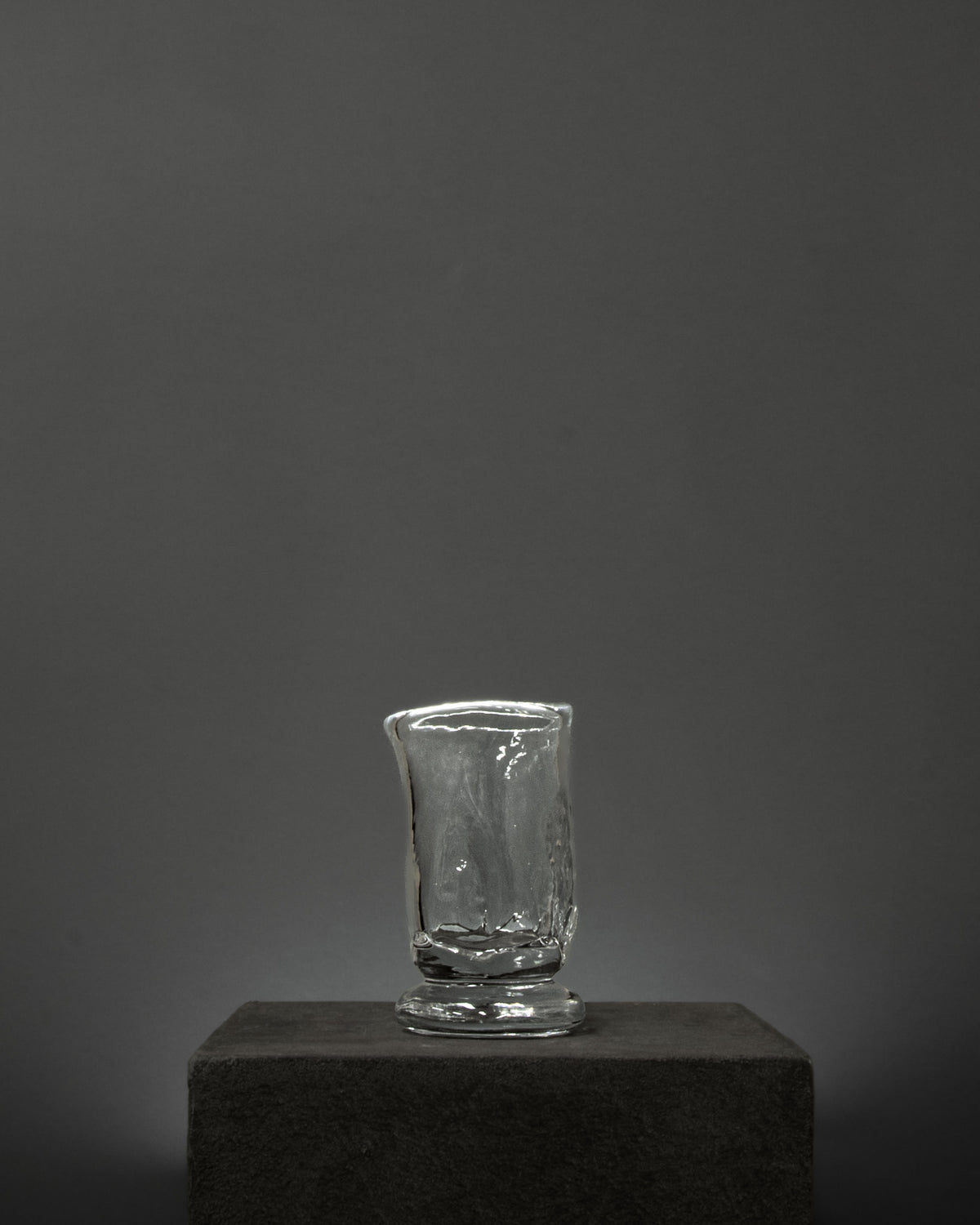 The Organic Shaped Table Glass by Szkło Studio rests on a dark, textured pedestal set against a plain gray background with soft lighting.