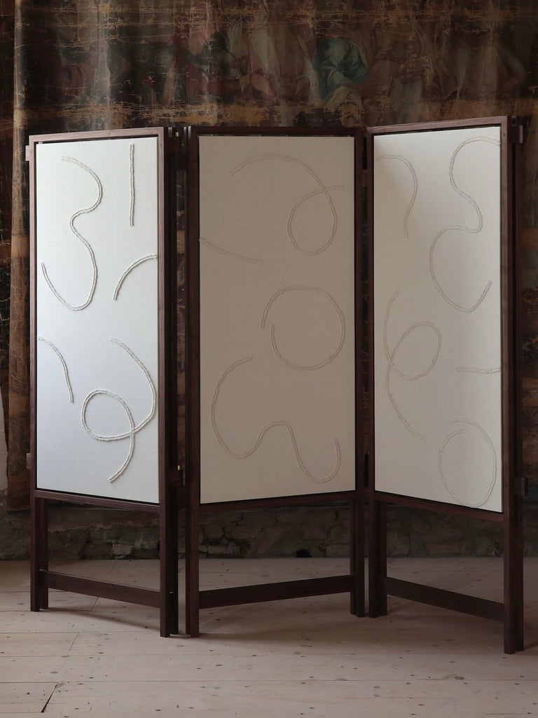 The Cavalletto Room Divider by Filippo Andrighetto features three white fabric panels with abstract curving line designs, set in a solid walnut frame—perfect as a stylish accent against textured walls and wooden floors.